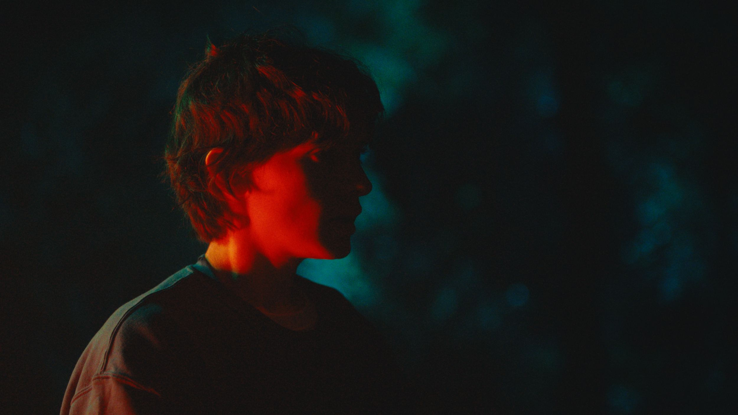 Profile of a young person with short, curly hair, wearing a dark jacket, illuminated by colorful, low lighting, with a dark, blurred background.