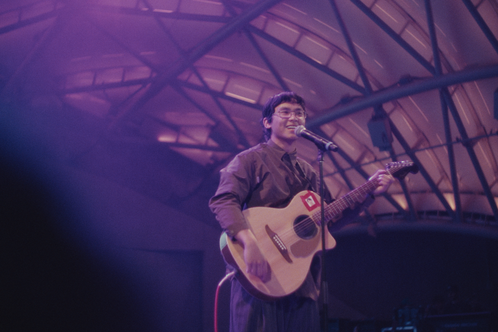 A young man with glasses, holding an acoustic guitar, standing on stage under purple lighting, singing into a microphone with a smile, with a large arched ceiling in the background.