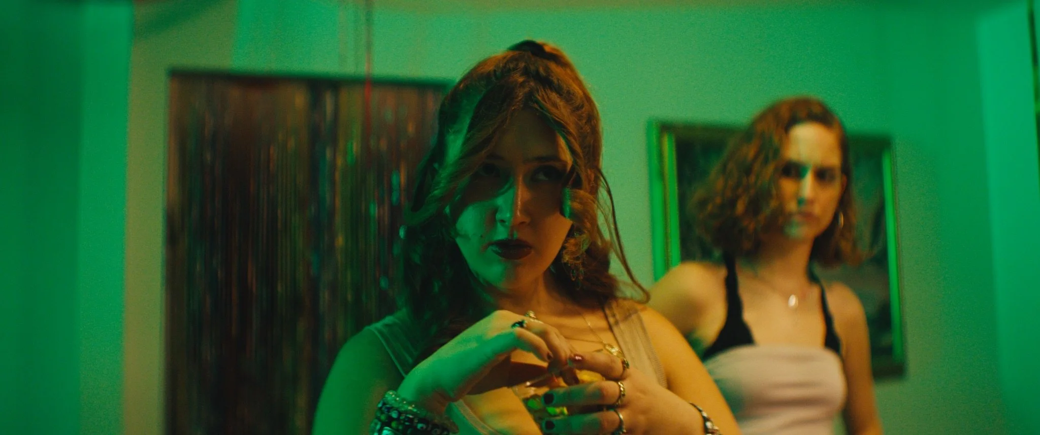 A woman with curly hair and dark lipstick looking into a mirror with another woman with similar hair in the background, both in low green lighting.