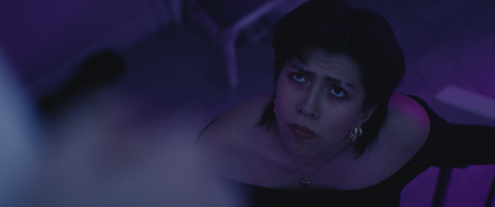 A woman with short dark hair, wearing earrings and a necklace, looks upward in a dimly lit room with purple lighting.