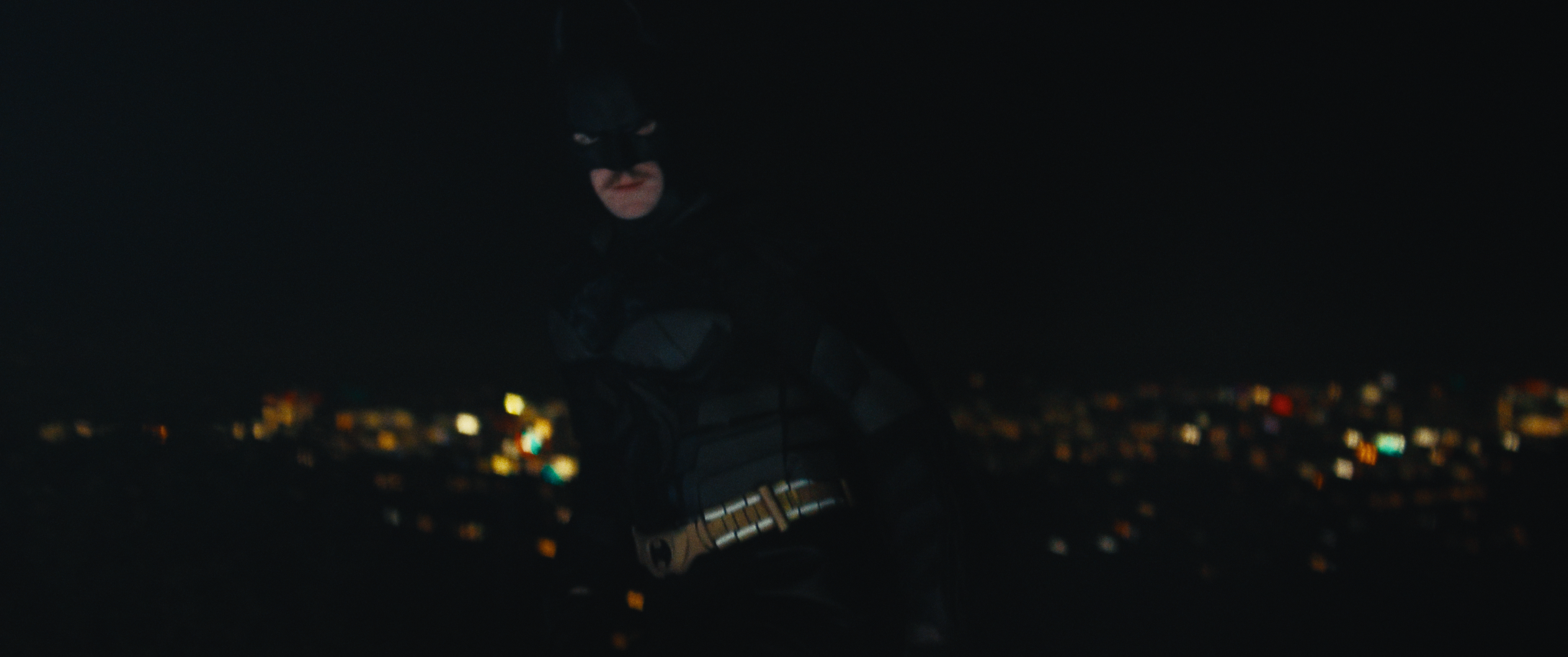 A person dressed as Batman standing on a rooftop at night with a cityscape in the background.