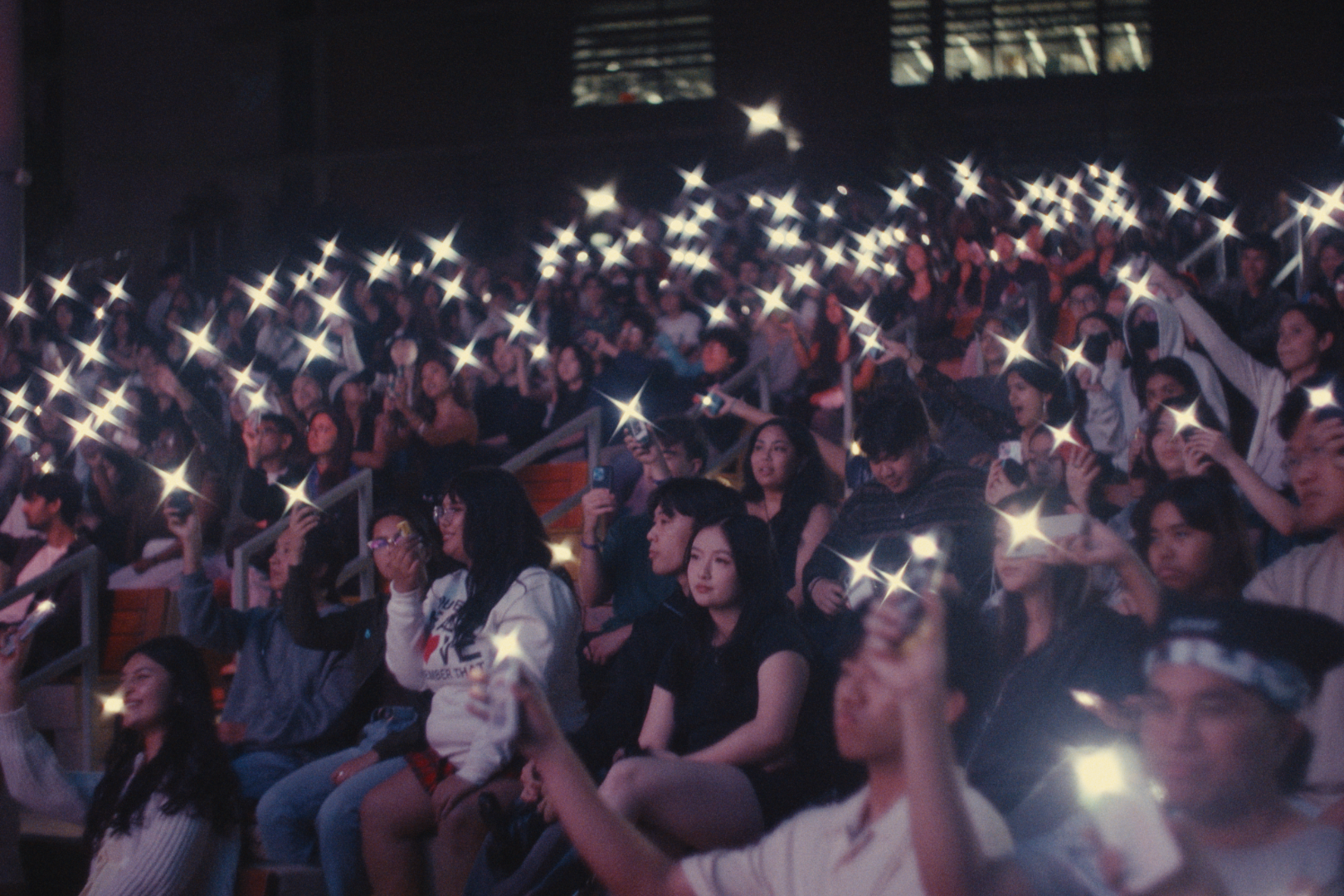 A large audience in a darkened indoor venue, with many people holding up their smartphones with flashlights on.