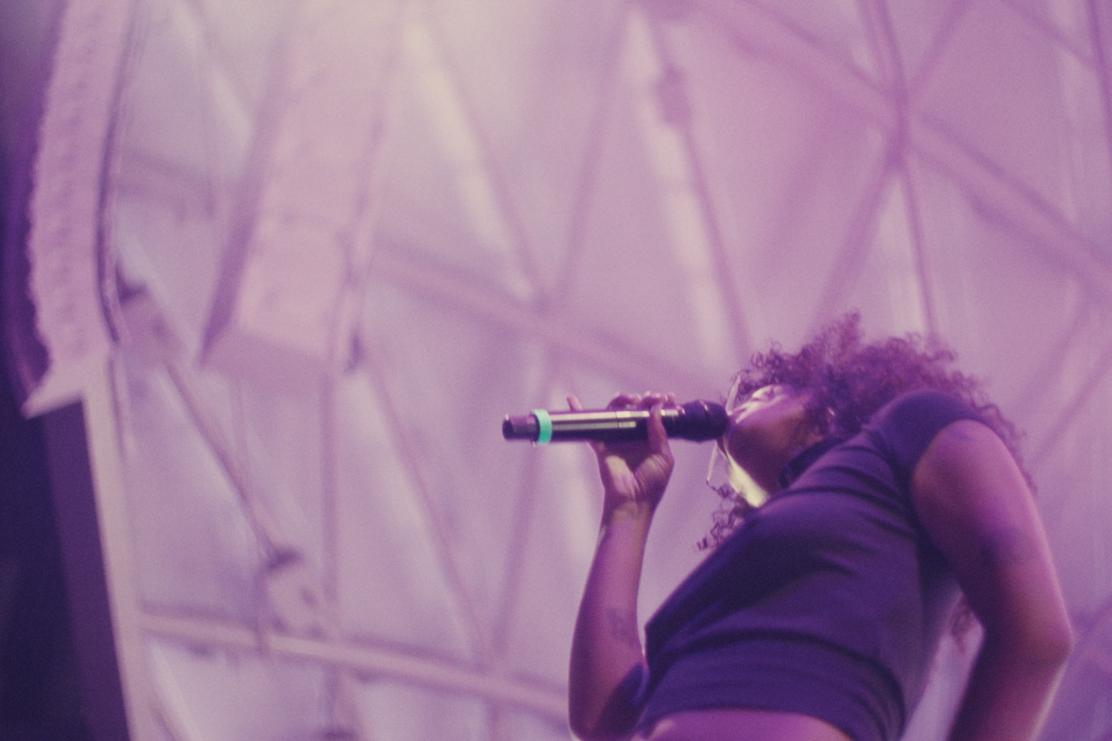 Female singer performing on stage, holding a microphone near her mouth, with purple stage lighting and abstract background.