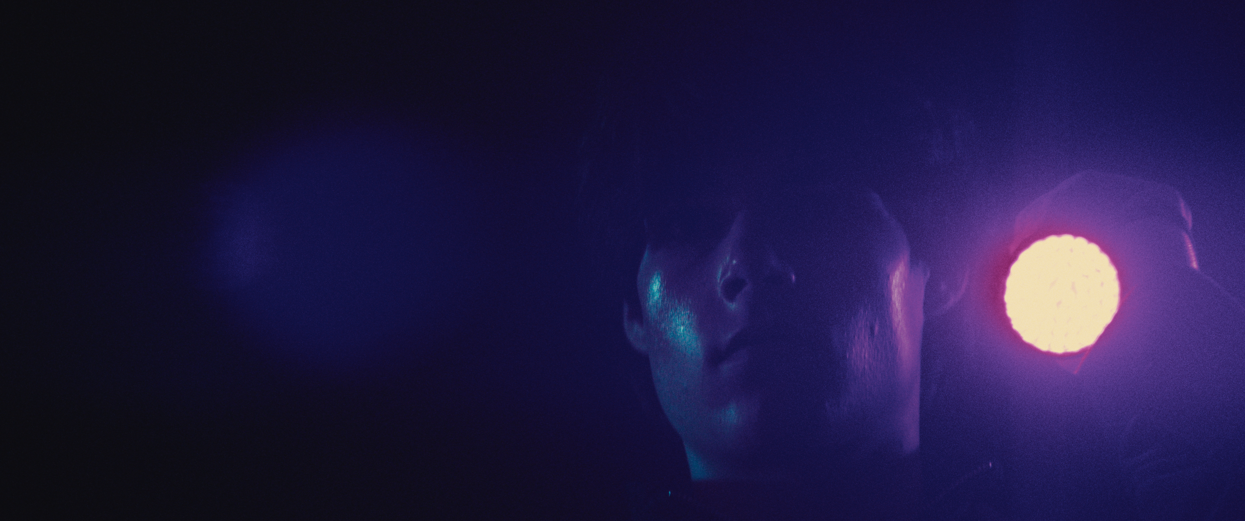 Person with face partially illuminated by purple and blue stage lights, dark background.