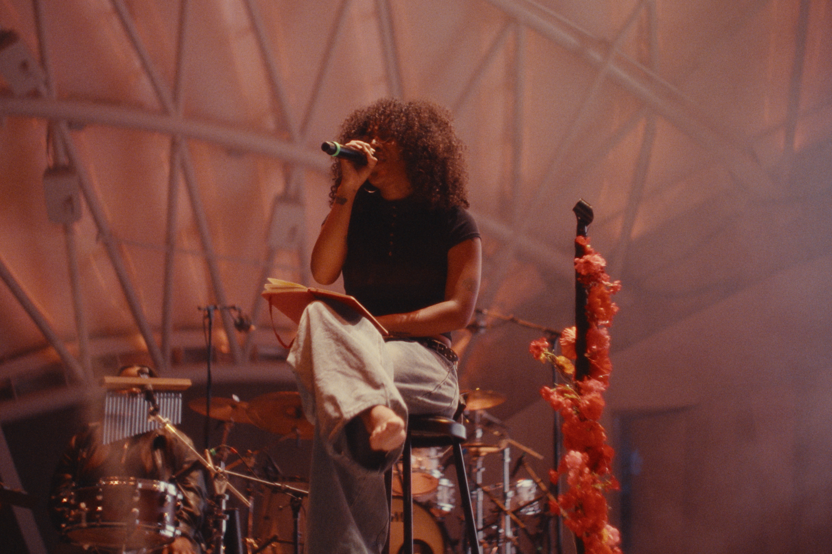 A woman with curly hair, wearing a black T-shirt and loose jeans, sitting on a stool with her right knee elevated, holding a microphone to her mouth and a notebook in her left hand, on stage with musical instruments and a microphone stand decorated w