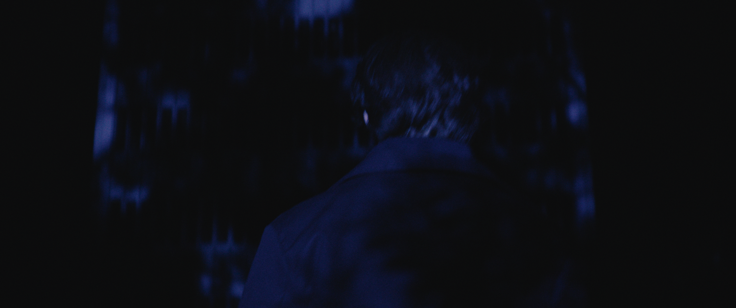 A person viewed from behind, walking through a dark or dimly lit environment with blue lighting.