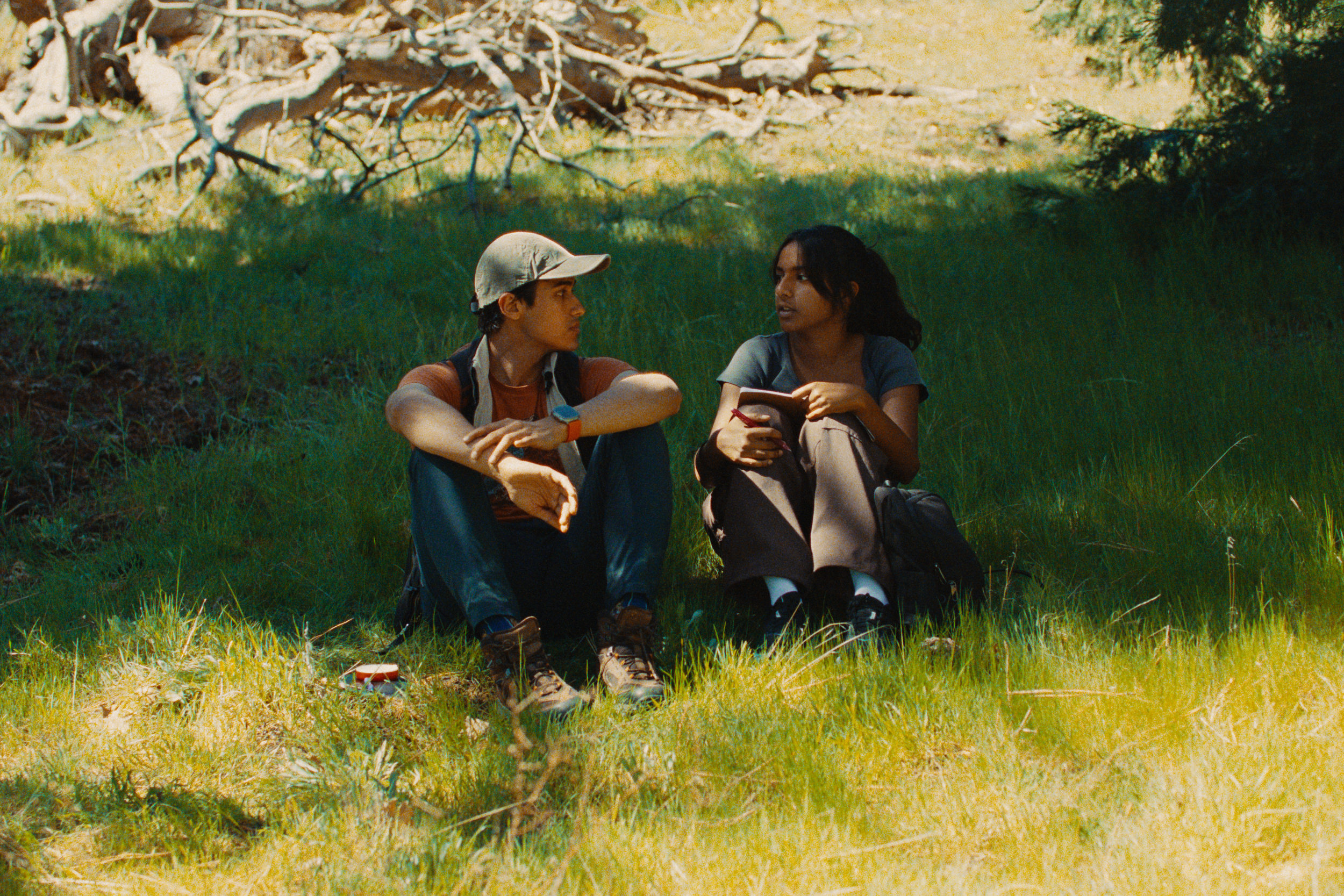 Two young women sitting on grass in a forested area, engaged in conversation. One is wearing a cap, and they both have backpacks and are dressed casually for outdoor activity.