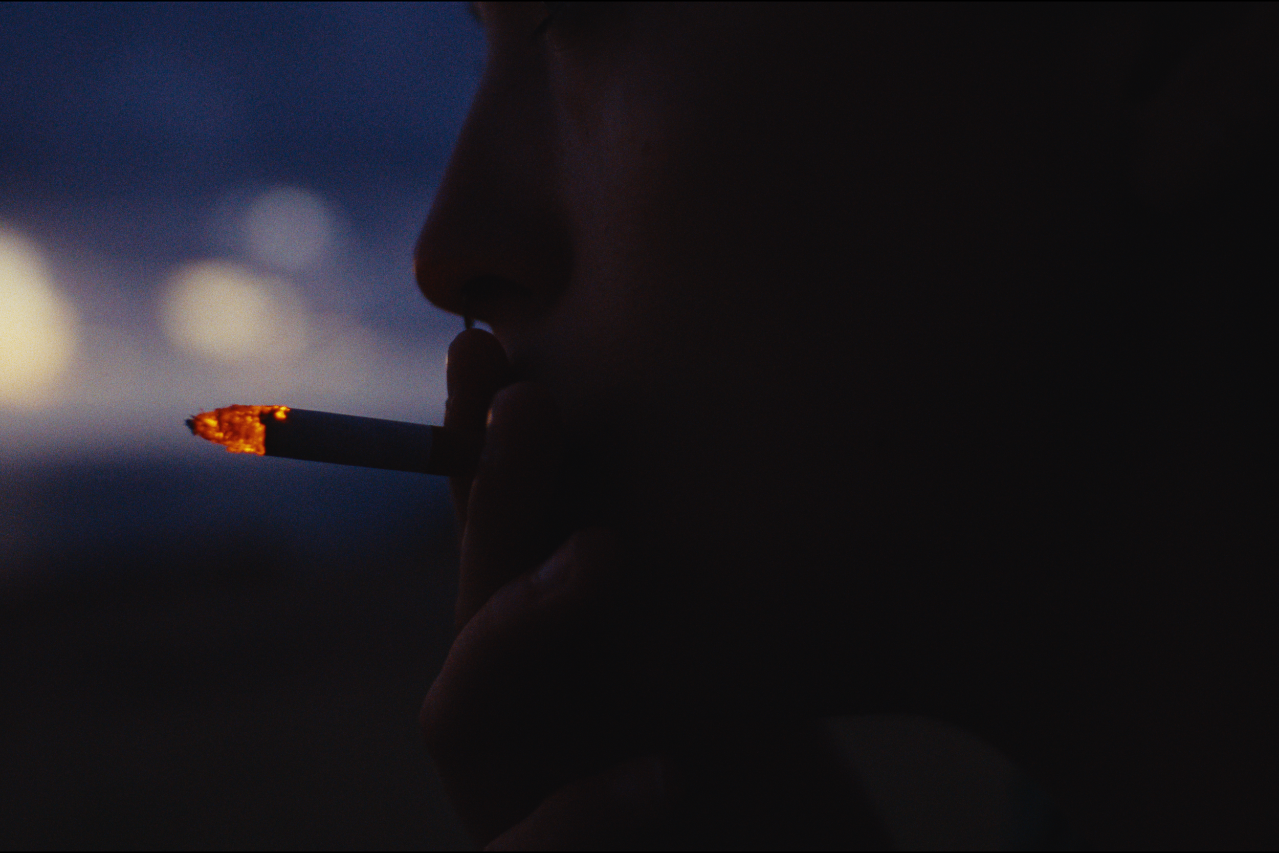 A person smoking a cigarette outdoors during dusk or evening, with a blurred background in shades of blue and orange.