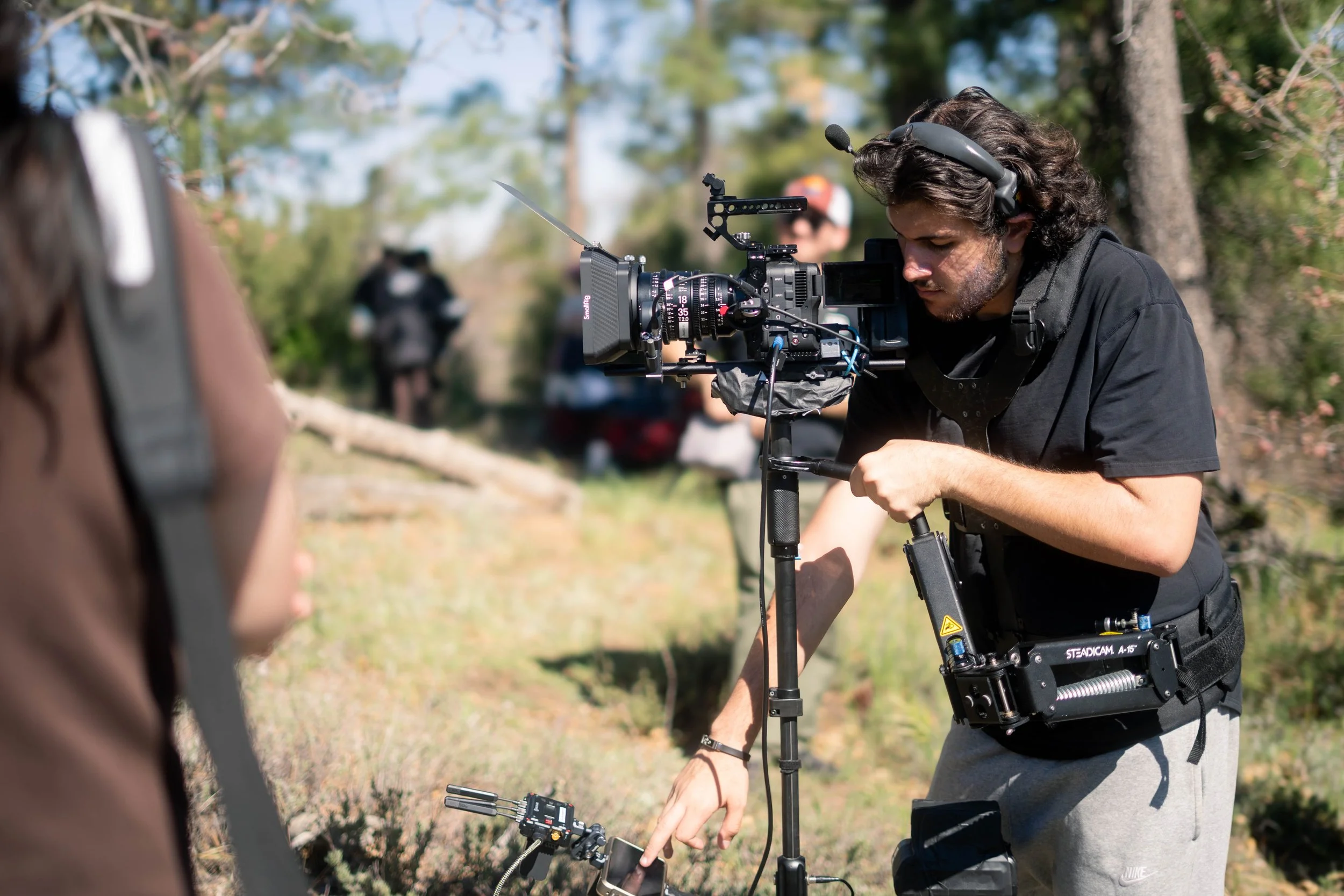 A man operating a professional camera with a shoulder rig outdoors, with two people in the background and trees around.