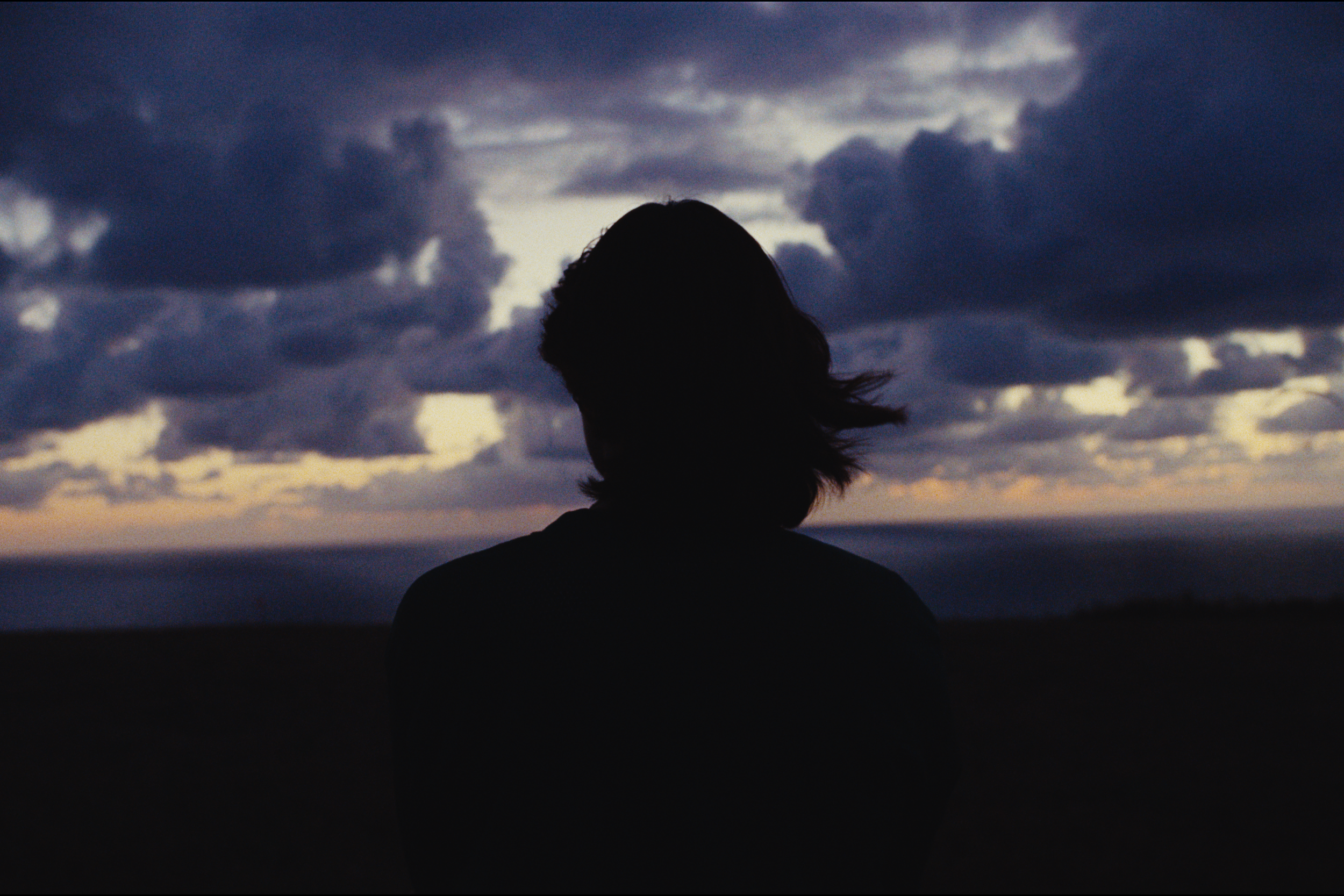 Silhouette of a person with long hair standing outdoors at dusk, clouds overhead, and a faint horizon glow in the background.