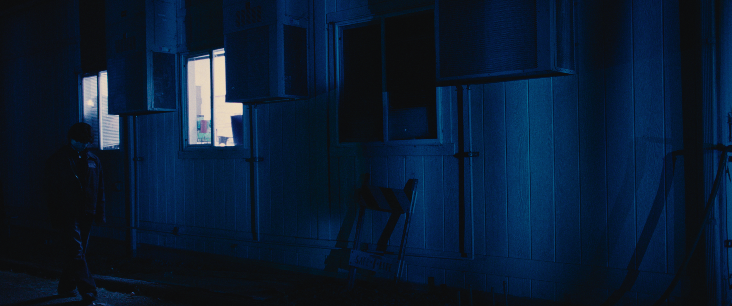 A person walking outside a building at night, with several open windows and some equipment on the ground, including a safety sign.
