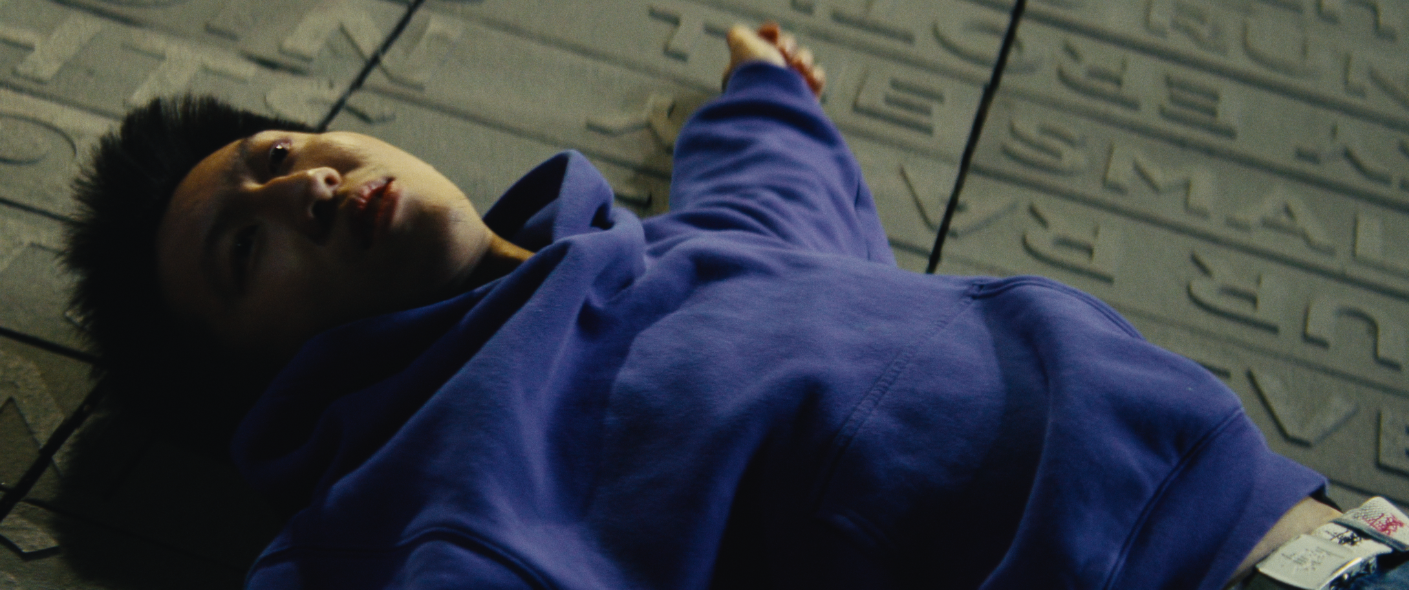 A young man lying on a tiled floor, looking up, wearing a blue hoodie.