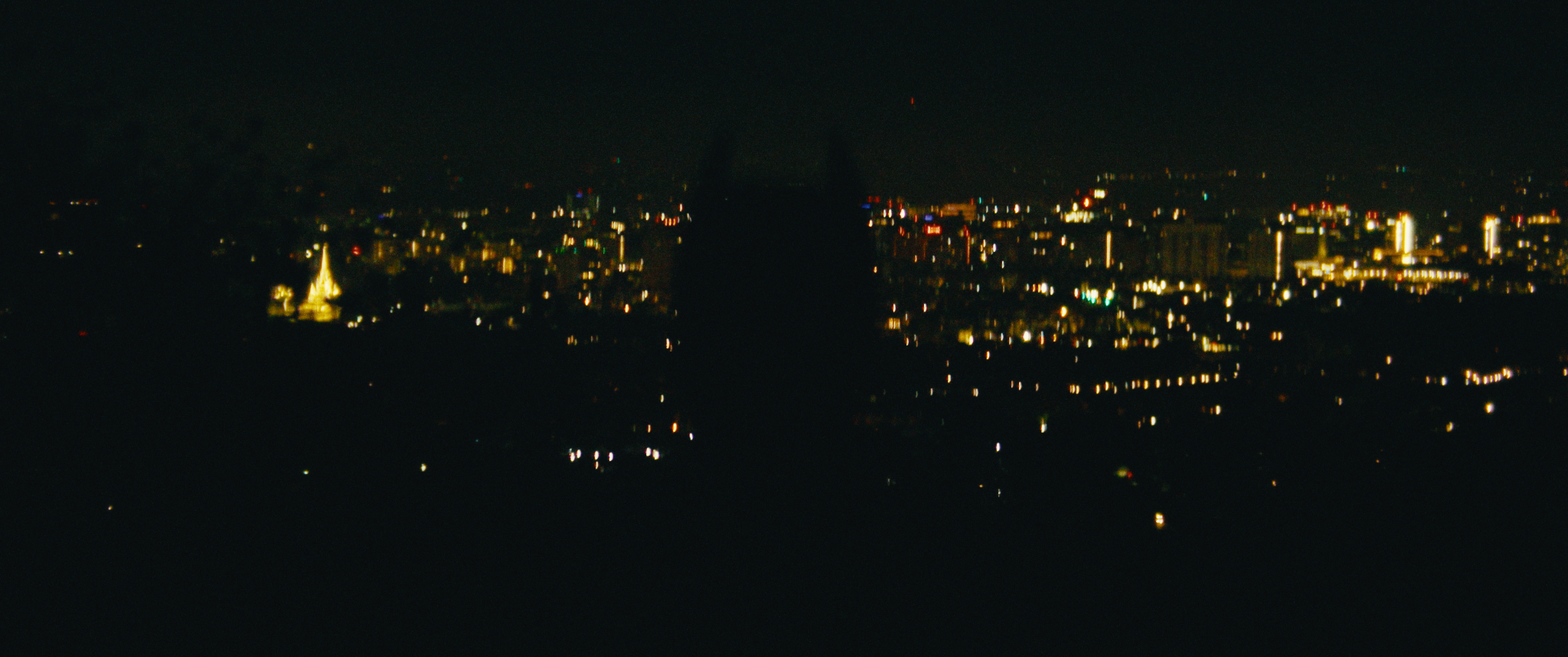 Nighttime view of a city skyline with buildings and skyscrapers illuminated, seen from a distance against a dark sky.