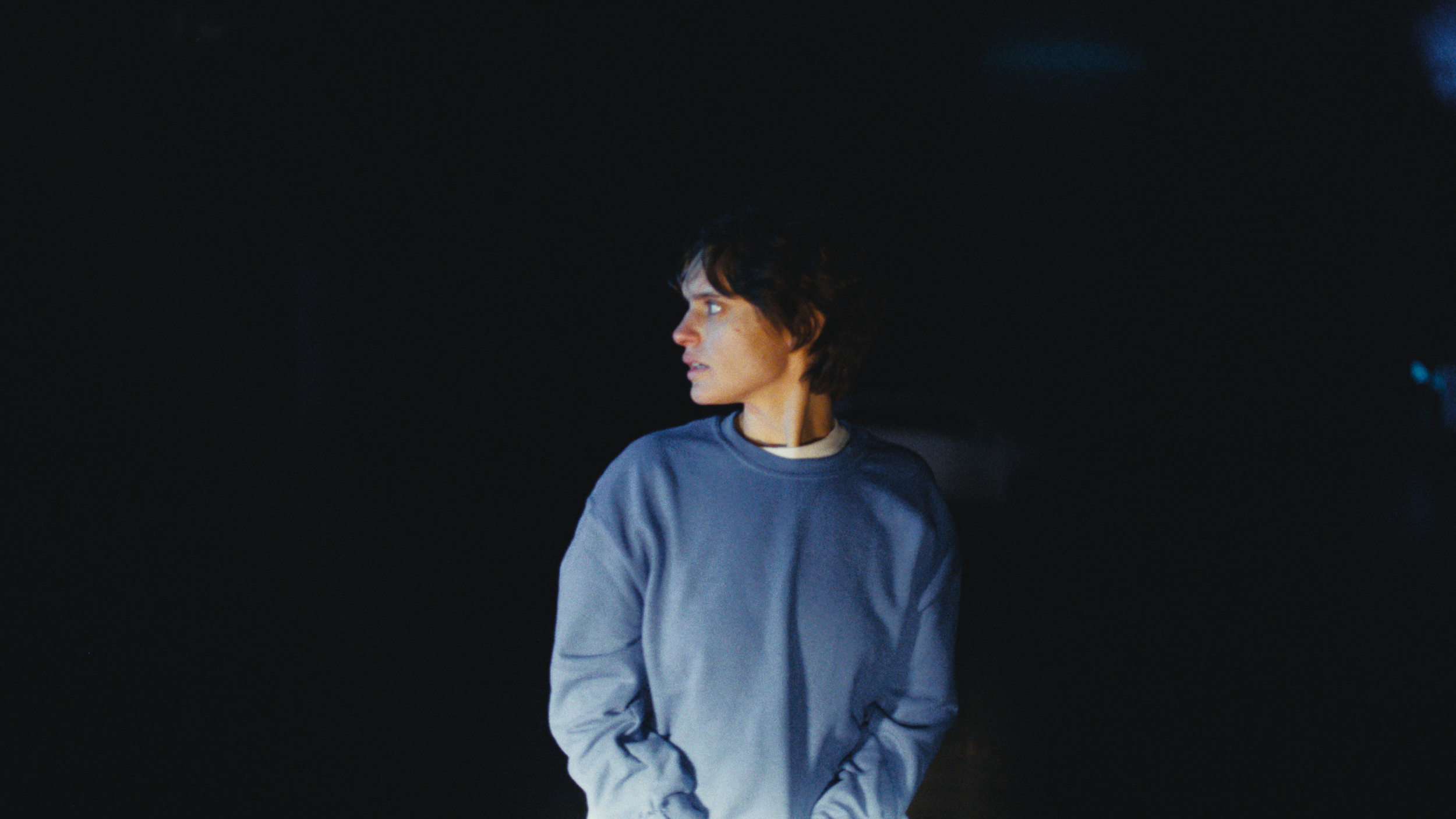 A person with short, dark, slightly curly hair wearing a blue sweatshirt standing against a dark background, looking to the side.