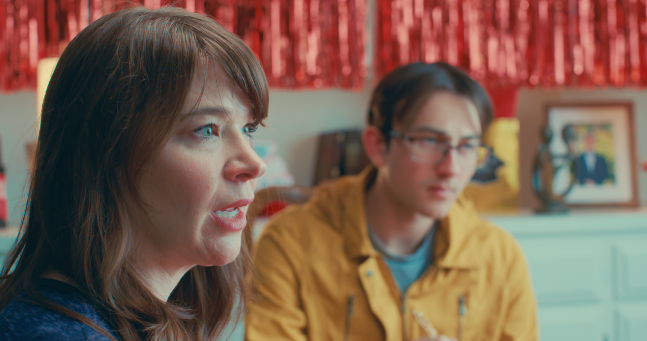 A woman with brown hair and blue eyes is talking, and a man with glasses and brown hair is sitting beside her, wearing a yellow jacket, in a room decorated with red curtains and framed photographs.