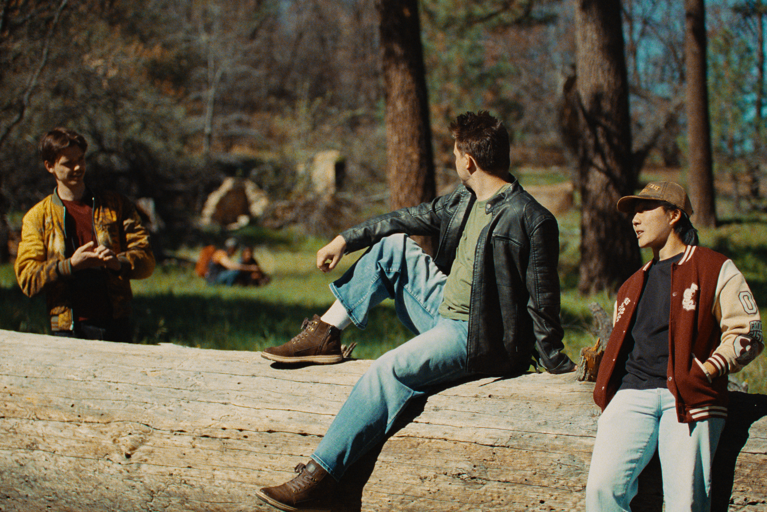 Three young men in casual clothing sitting and standing on a log in a forested park. One is standing, smiling and talking; another is sitting with his legs outstretched, facing sideways; the third is sitting and facing forward, wearing a baseball cap