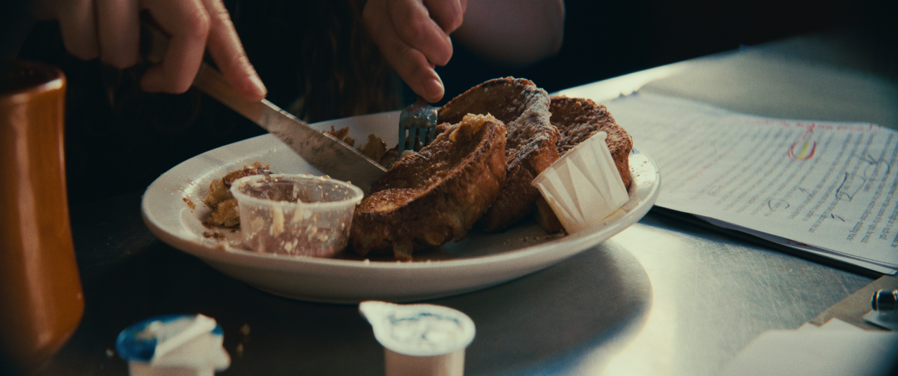 A person is cutting into French toast with syrup, on a white plate with a small container of syrup and paper muffin liner, on a table with papers and a pen.