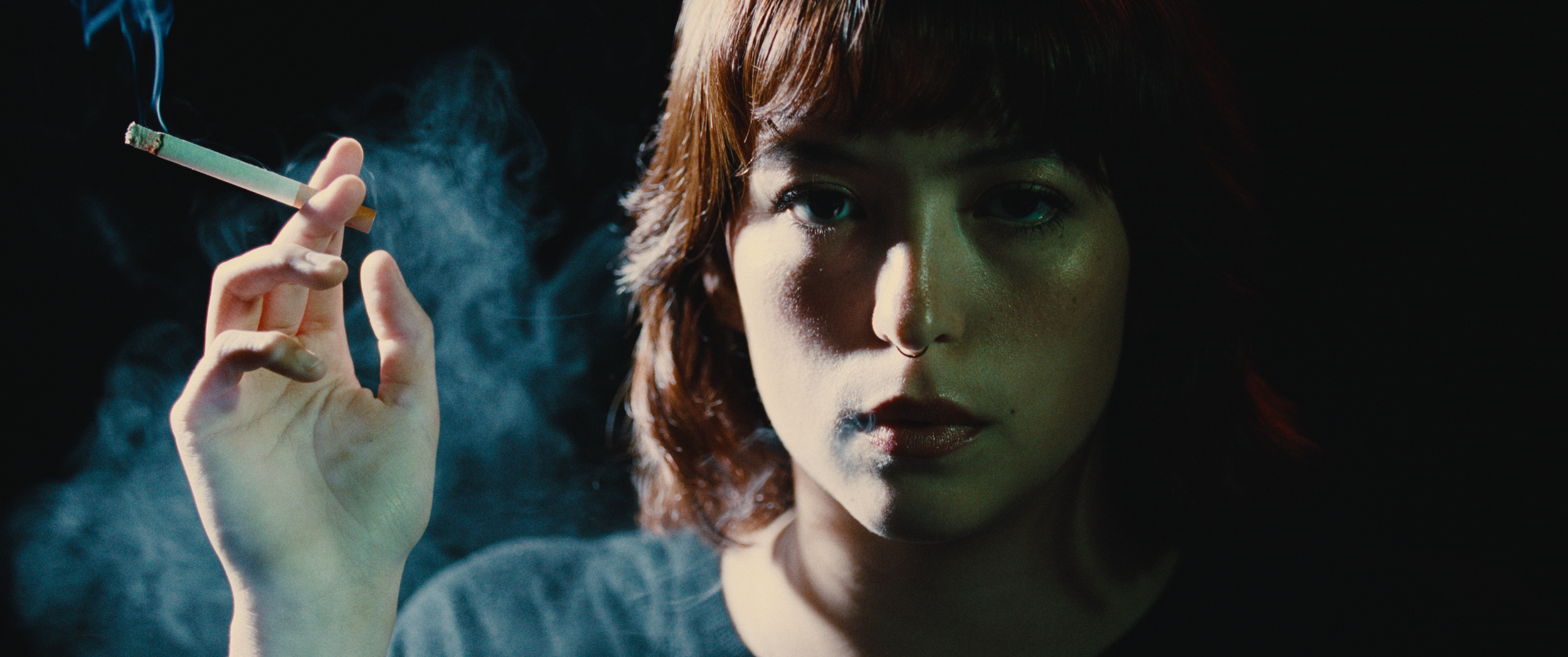 A young woman with dark hair and a septum piercing holds a lit cigarette in her hand, with smoke swirling around her face in a dark setting.