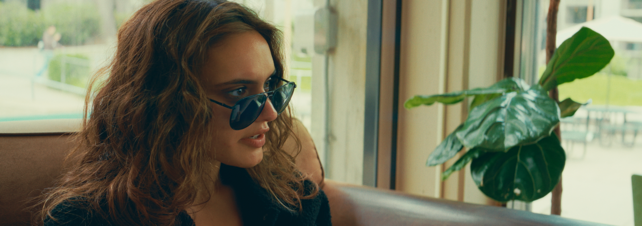 Woman with curly hair and sunglasses sitting indoors near a large window, with a large green plant beside her.
