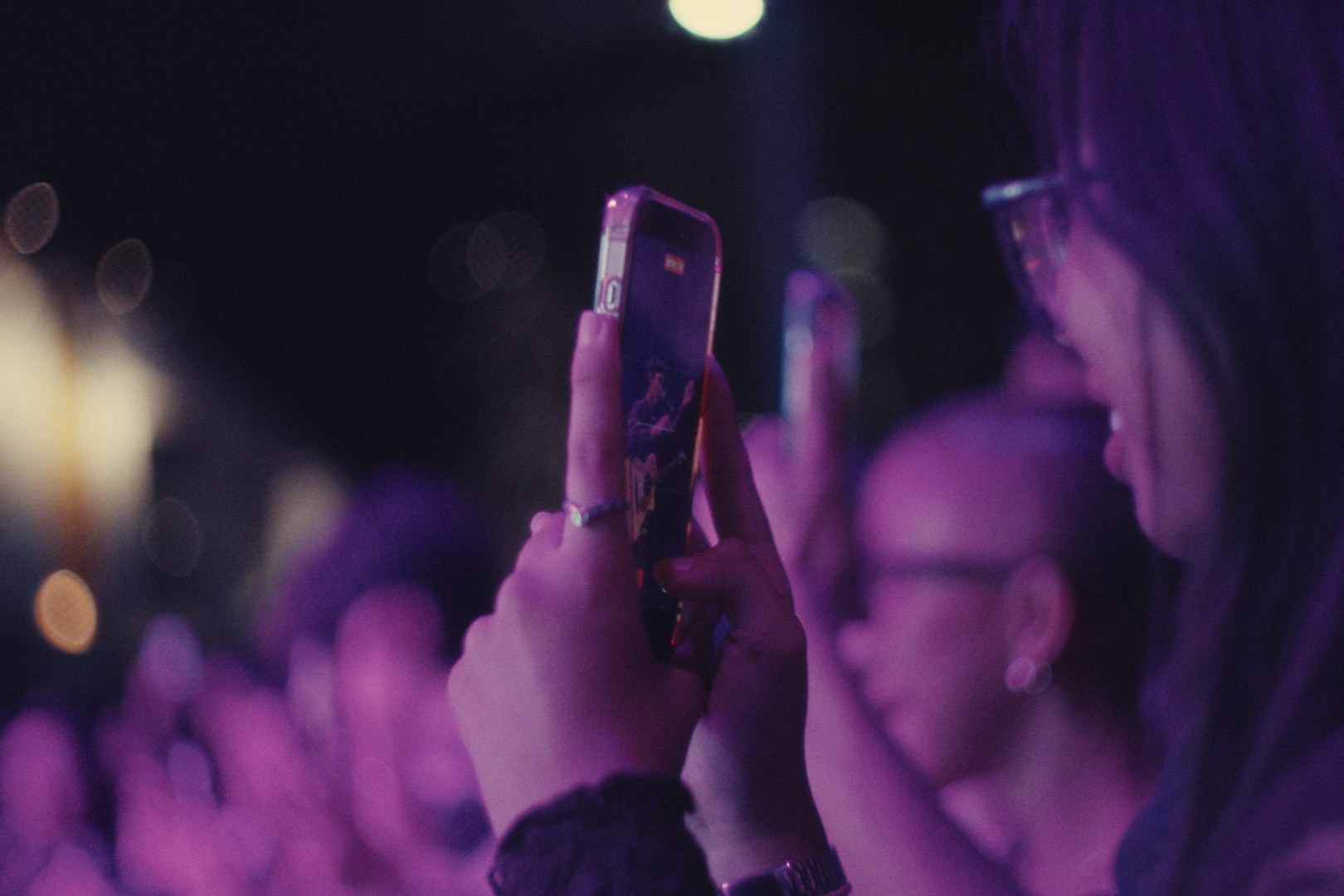 A woman with glasses taking a photo or video with her smartphone at a nighttime event, surrounded by other people, with city lights and bokeh in the background.