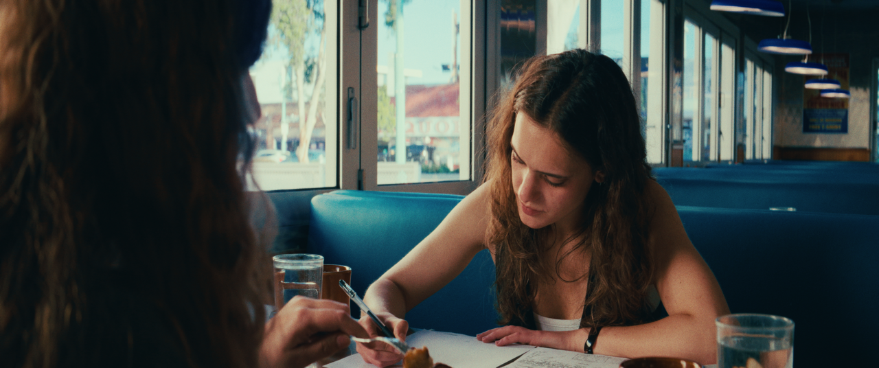 A woman with wavy brown hair sitting at a table in a restaurant, looking down at a notebook, with a glass of water in front of her and another person partially visible on the left.