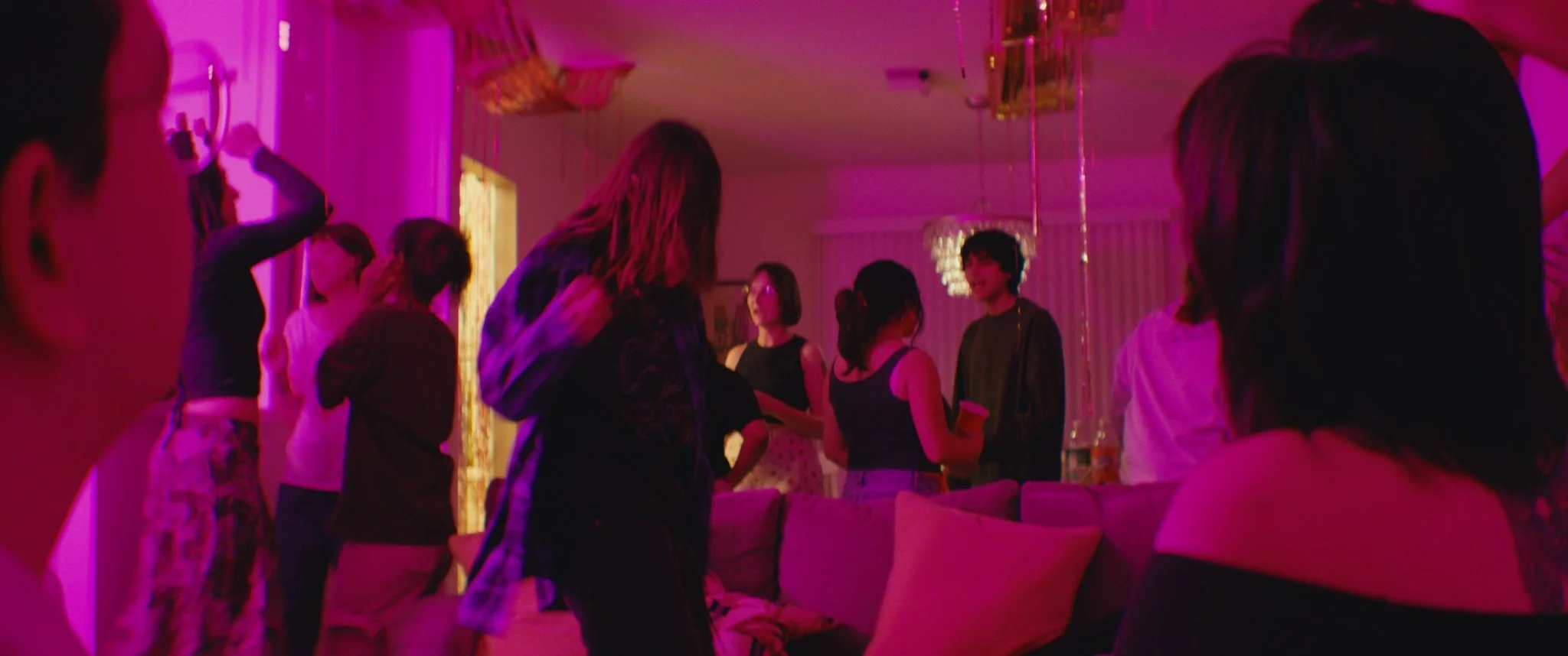 A group of people socializing and dancing at a party or gathering in a room lit with pink and purple lights.