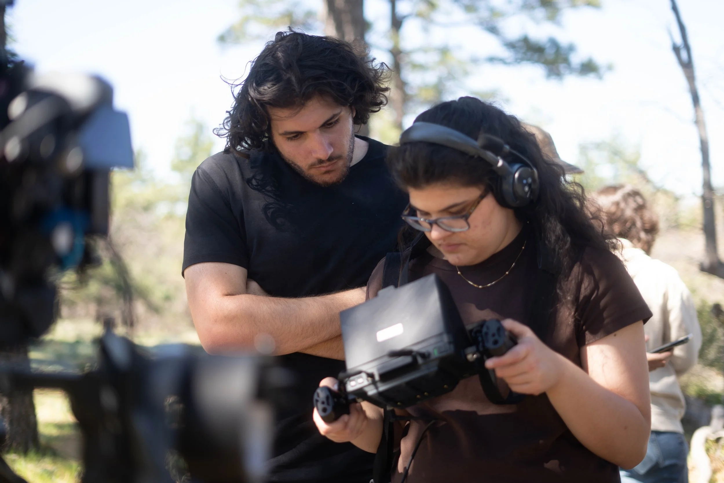 Two young women outdoors, one wearing headphones, examining a handheld device while a camera is set up in front of them.