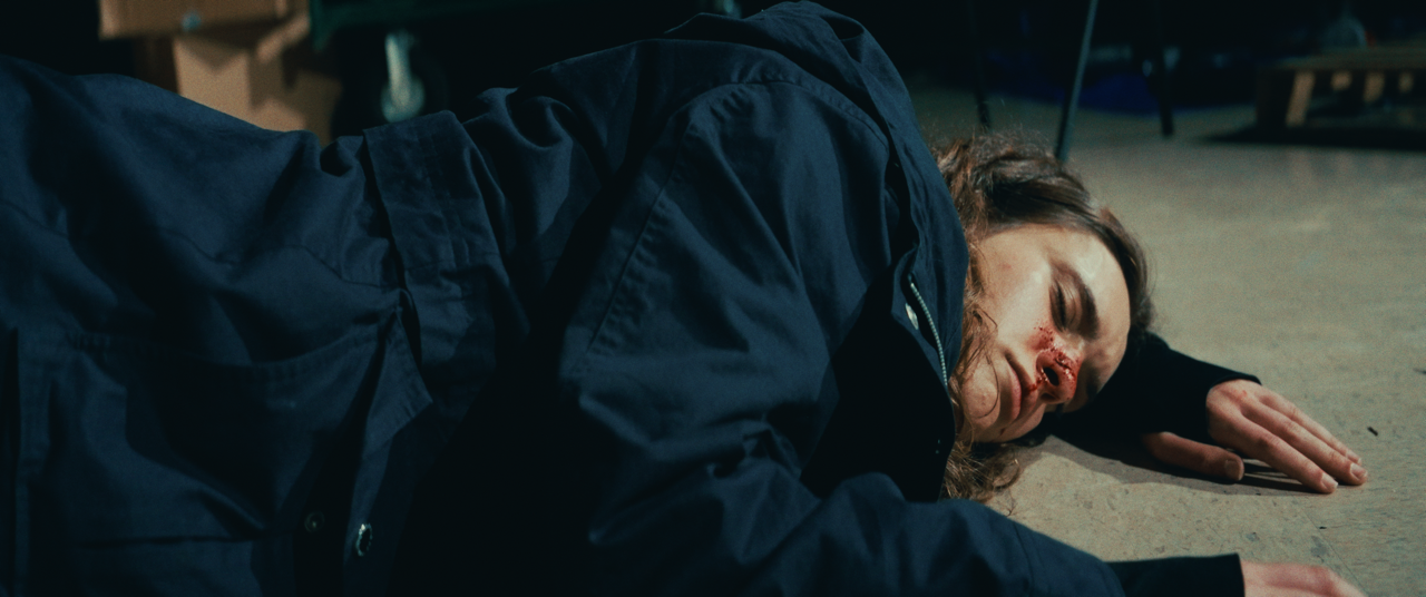 A woman lies on the floor with a nosebleed, appearing to be unconscious or injured, in a dimly lit room.