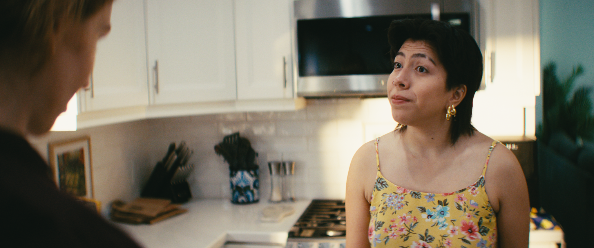 A woman with short dark hair wearing a yellow floral dress and gold hoop earrings talking to a person with blond hair in a kitchen with white cabinets and a stainless steel microwave.