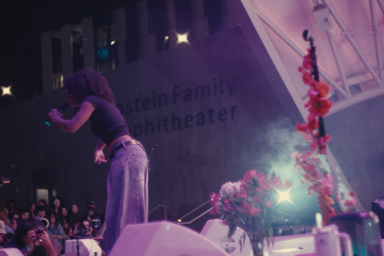 A performer with curly hair on stage at the Stein Family Amphitheater during an evening event, holding a microphone while standing in front of an audience with photographers capturing the moment. A floral arrangement is visible in the foreground.