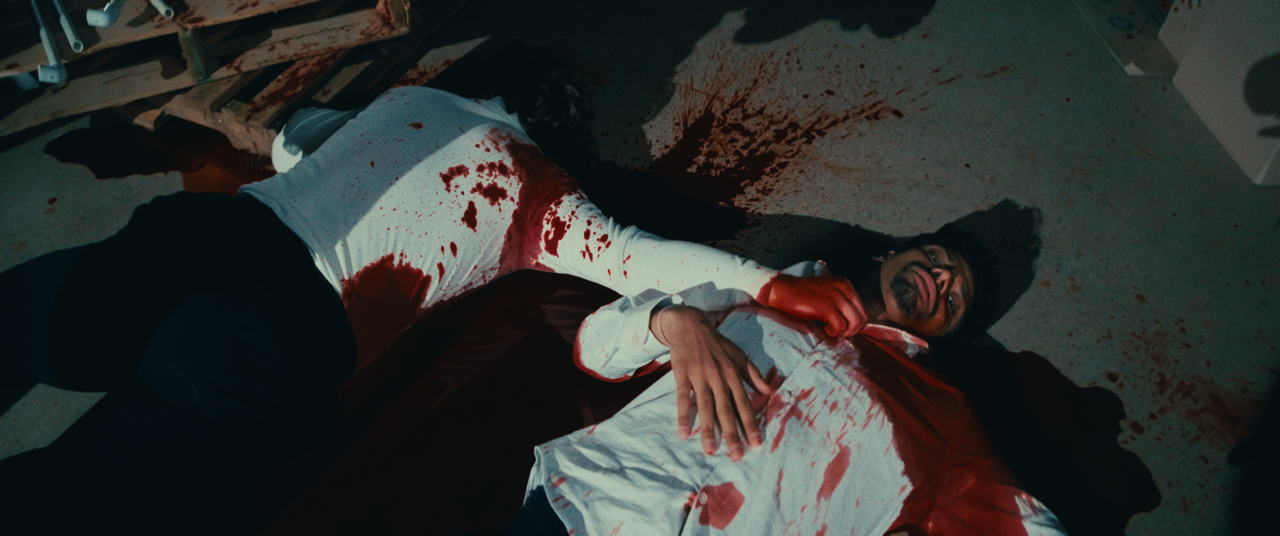 A man lying on the floor covered in blood, with blood spatters around him, wearing a white shirt and red gloves, appearing to be in a dramatic or violent scene.