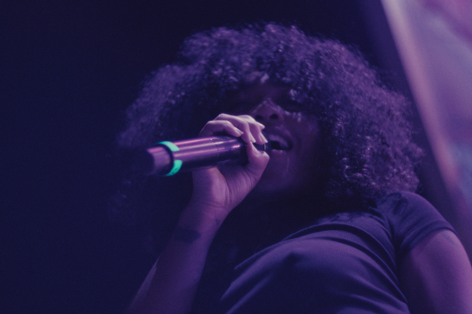 Woman with large curly hair singing into a microphone in a dark setting with purple lighting.