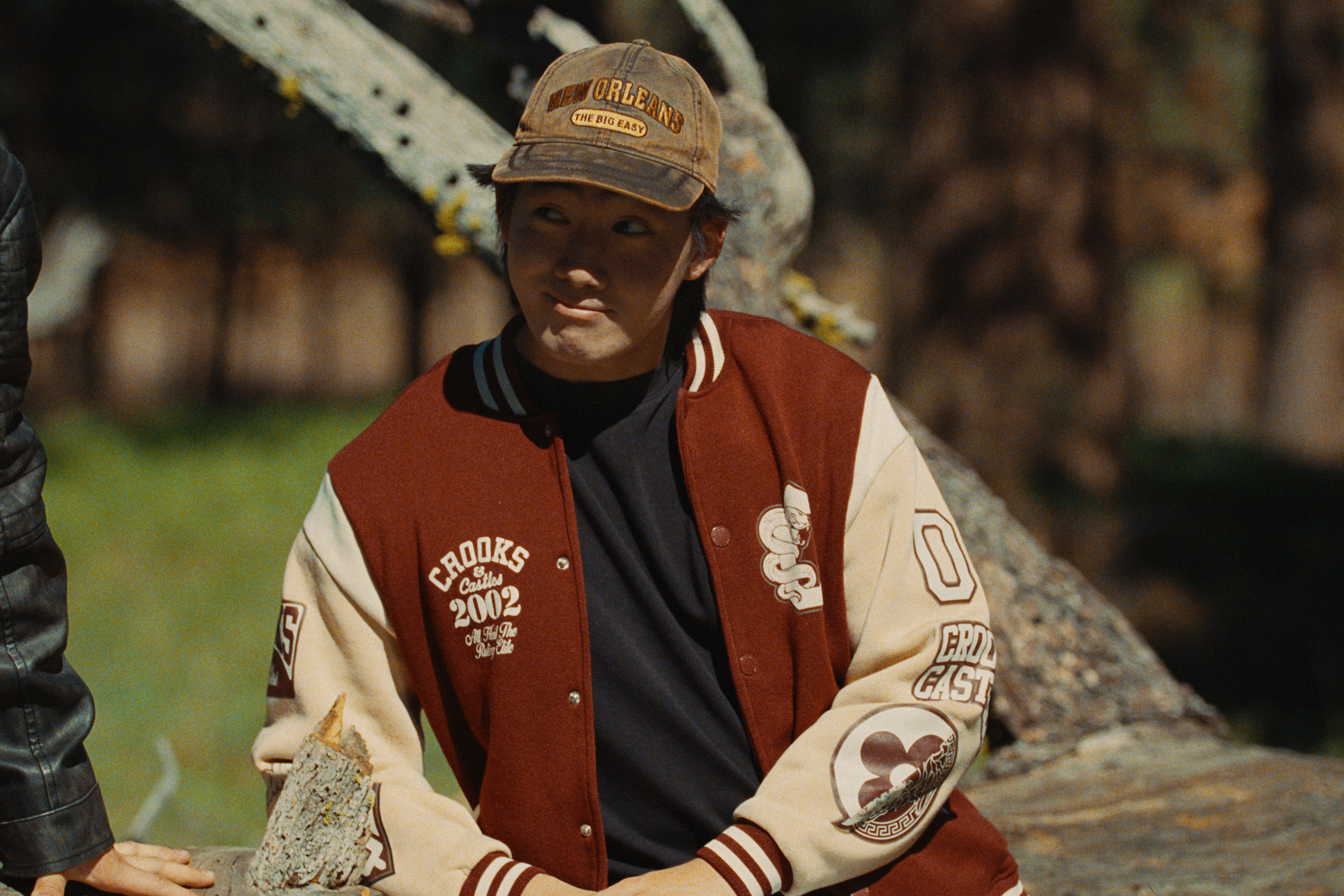 A young boy with black hair wearing a beige and maroon varsity jacket and a brown baseball cap with the words 'New Orleans' and 'The Big Easy' on it. He is outdoors, next to a tree, holding a piece of wood or bark.