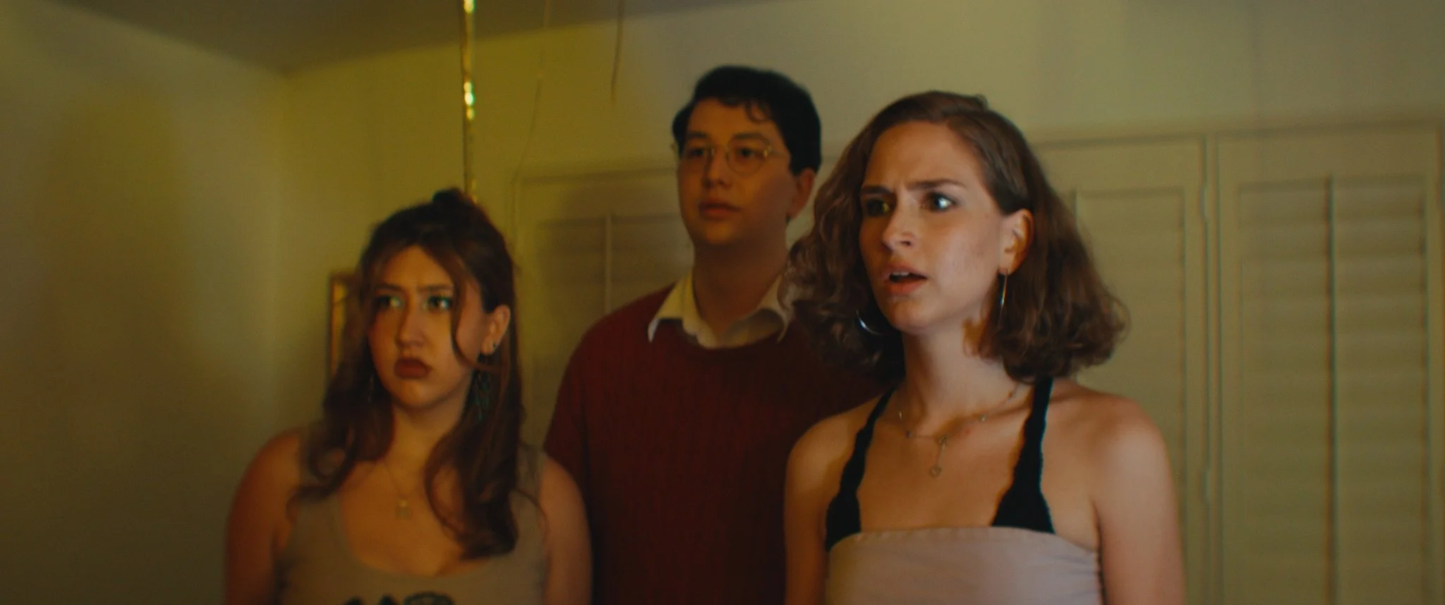 Three young adults standing close together in a dimly lit room, looking with expressions of surprise or concern.