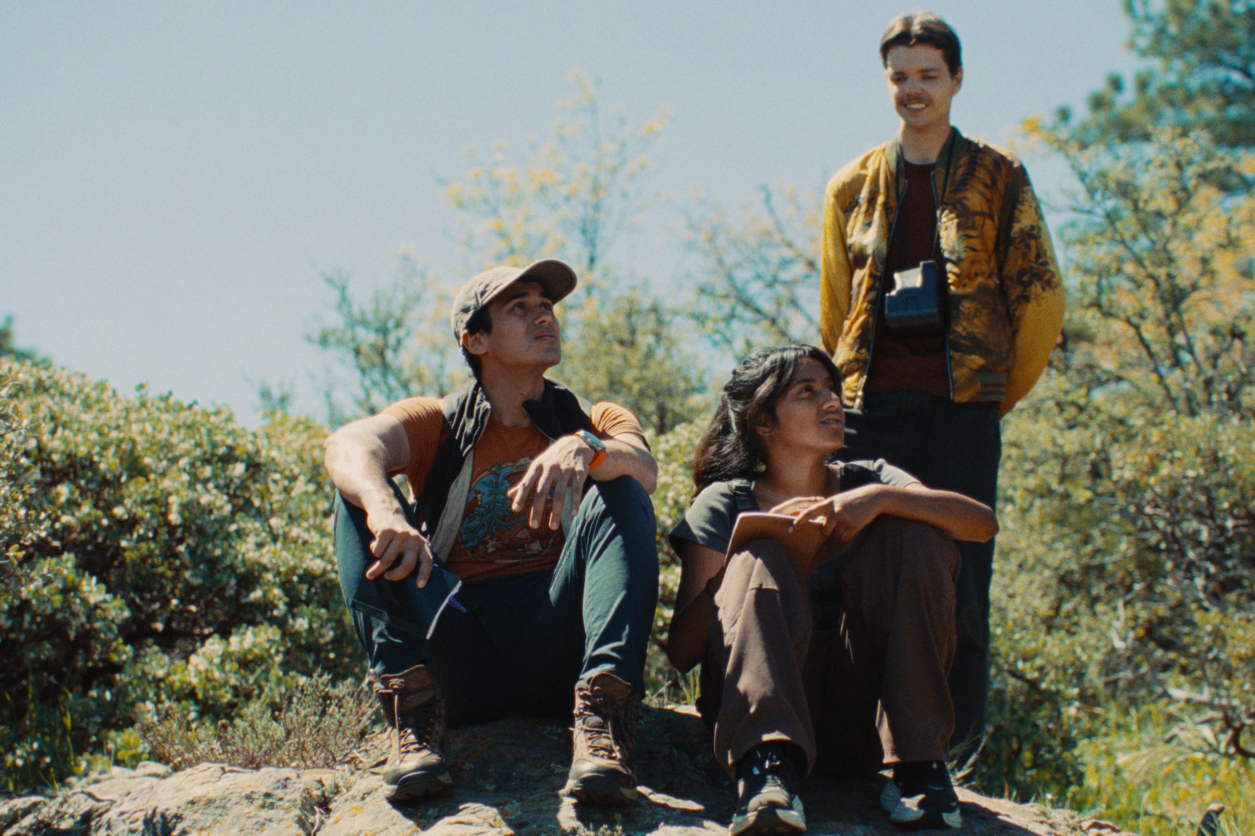 Three hikers sitting outdoors on a flat rock, two women and one man, with trees and shrubs in the background, one woman and the man are looking up and the other woman is looking ahead.