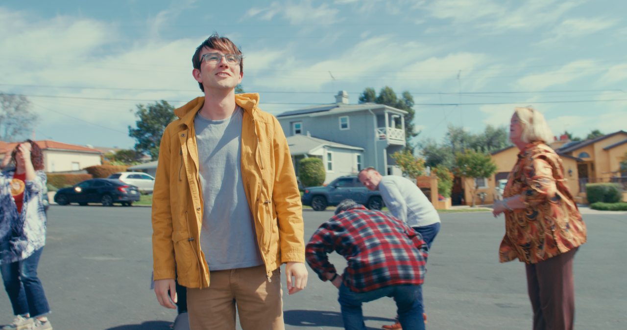 Young man in glasses and a yellow jacket standing outdoors in a neighborhood street on a sunny day, surrounded by other people.