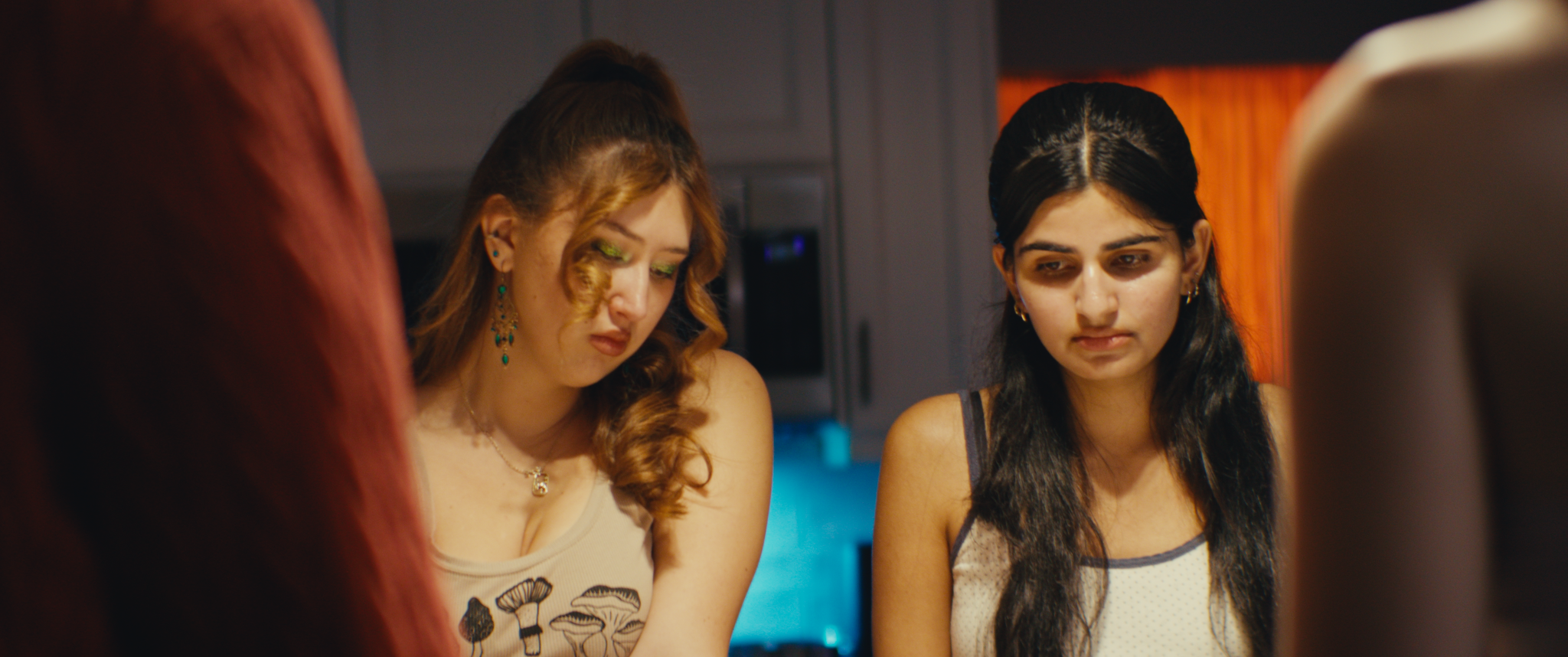 Two women standing close together indoors, with serious expressions, in a dimly lit room with blue and orange lighting.