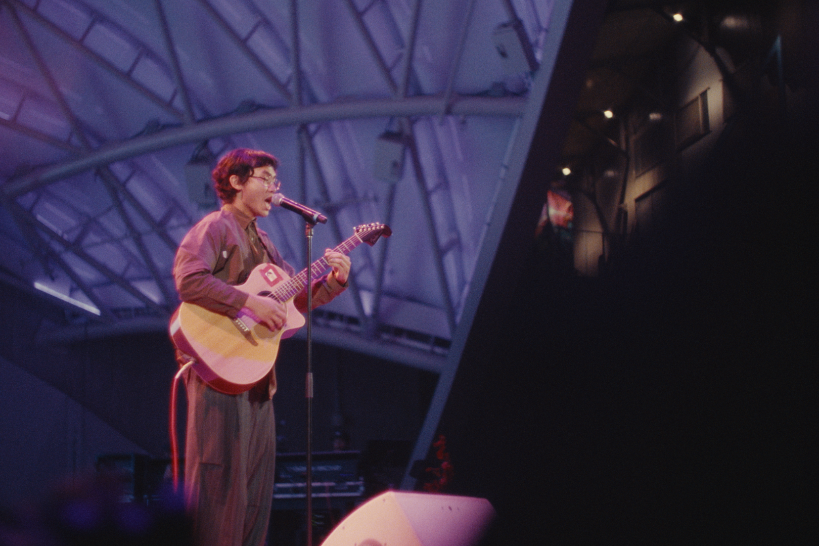 A person with glasses singing into a microphone while playing an acoustic guitar on stage, under purple lighting, with an architectural ceiling structure visible in the background.