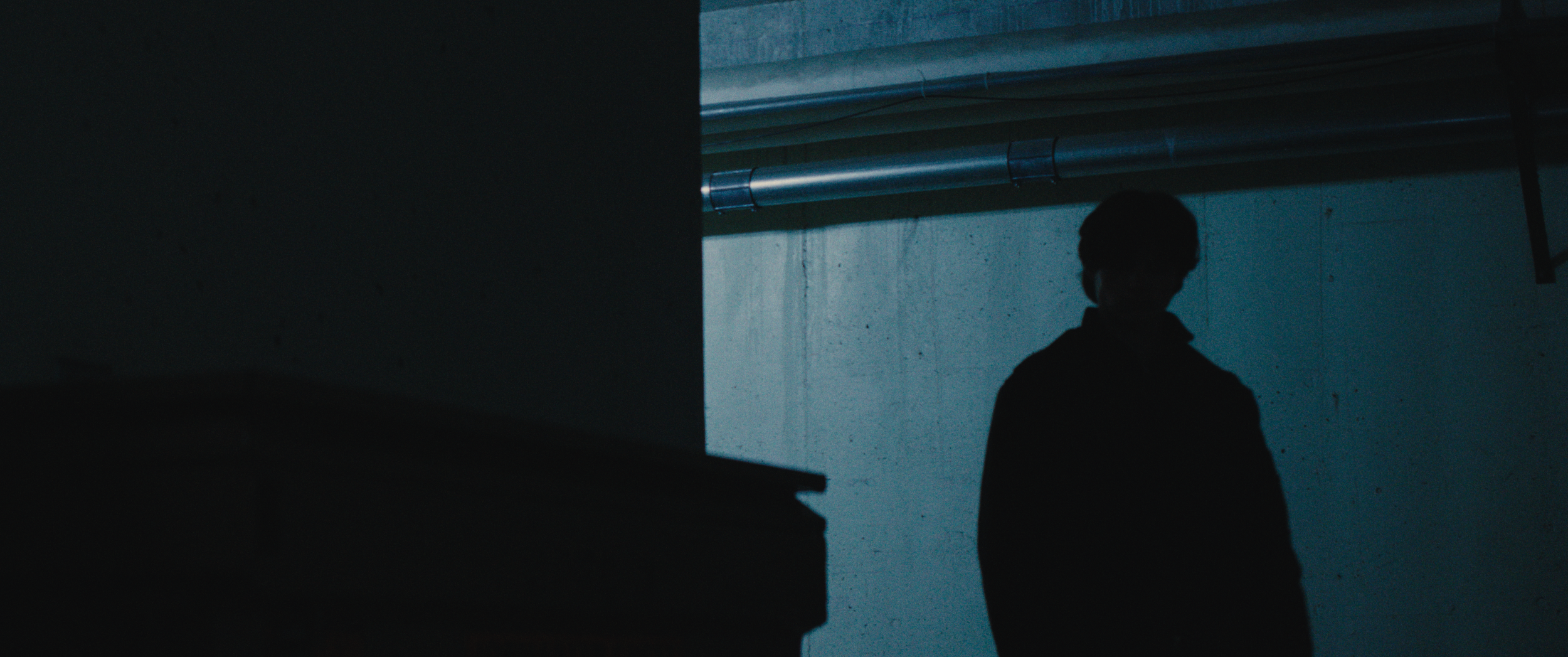 Silhouette of a person standing in a dimly lit concrete space with exposed pipes on the ceiling.
