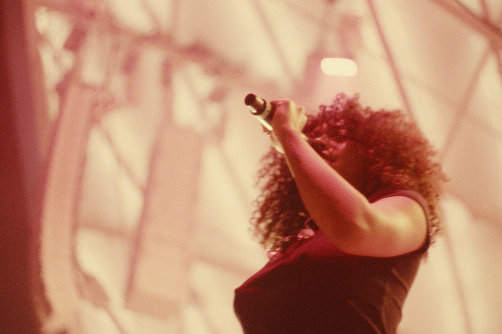 A person with curly hair holding a microphone, singing or speaking, against a decorated background with warm lighting.
