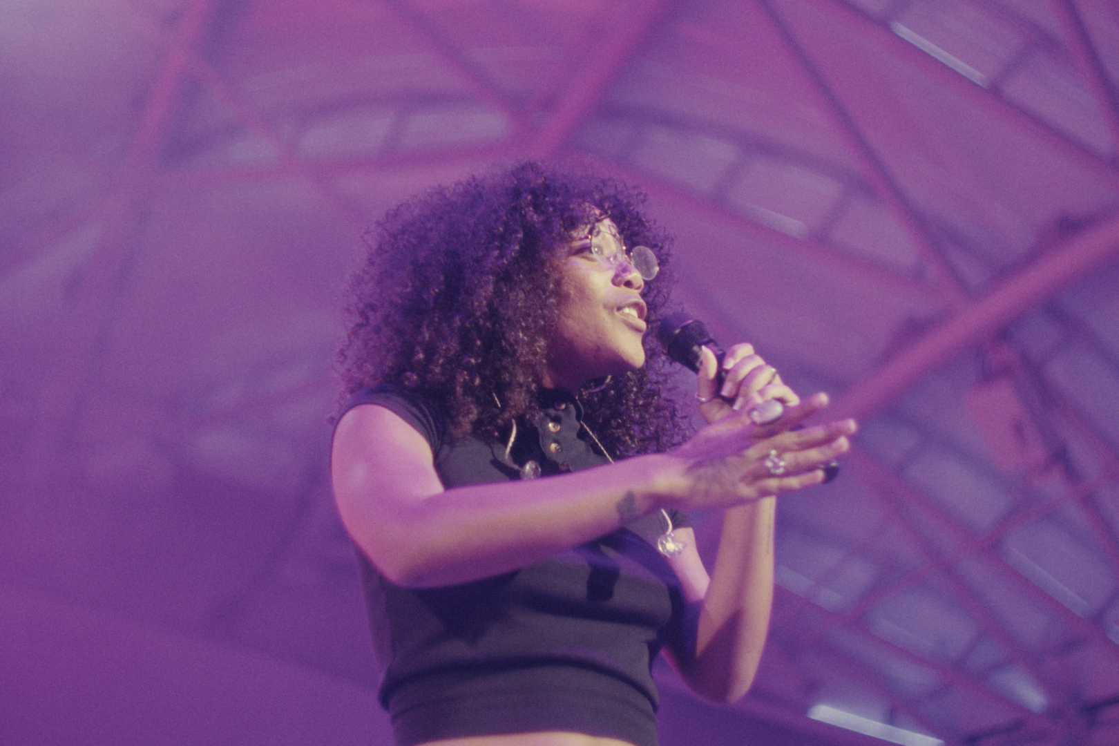 A woman with curly hair and glasses speaking into a microphone on a stage with purple neon lights.