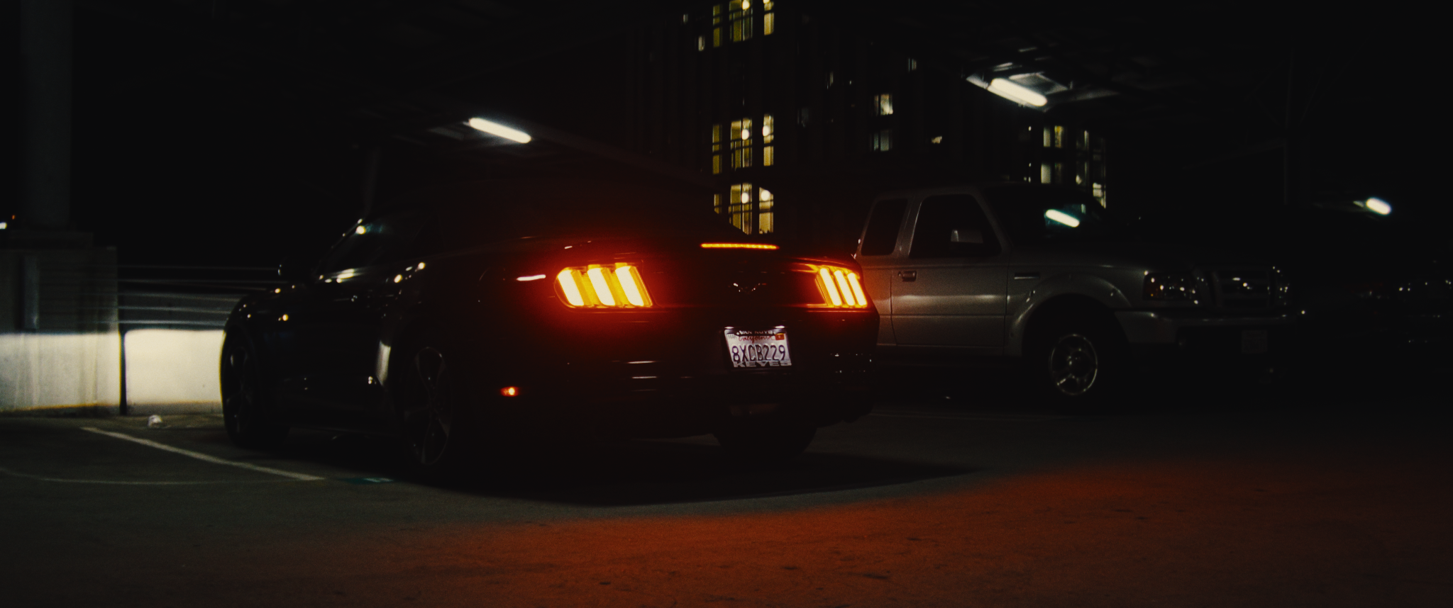 A black sports car with illuminated taillights parked in a dimly lit parking garage at night, with a gray pickup truck parked beside it.