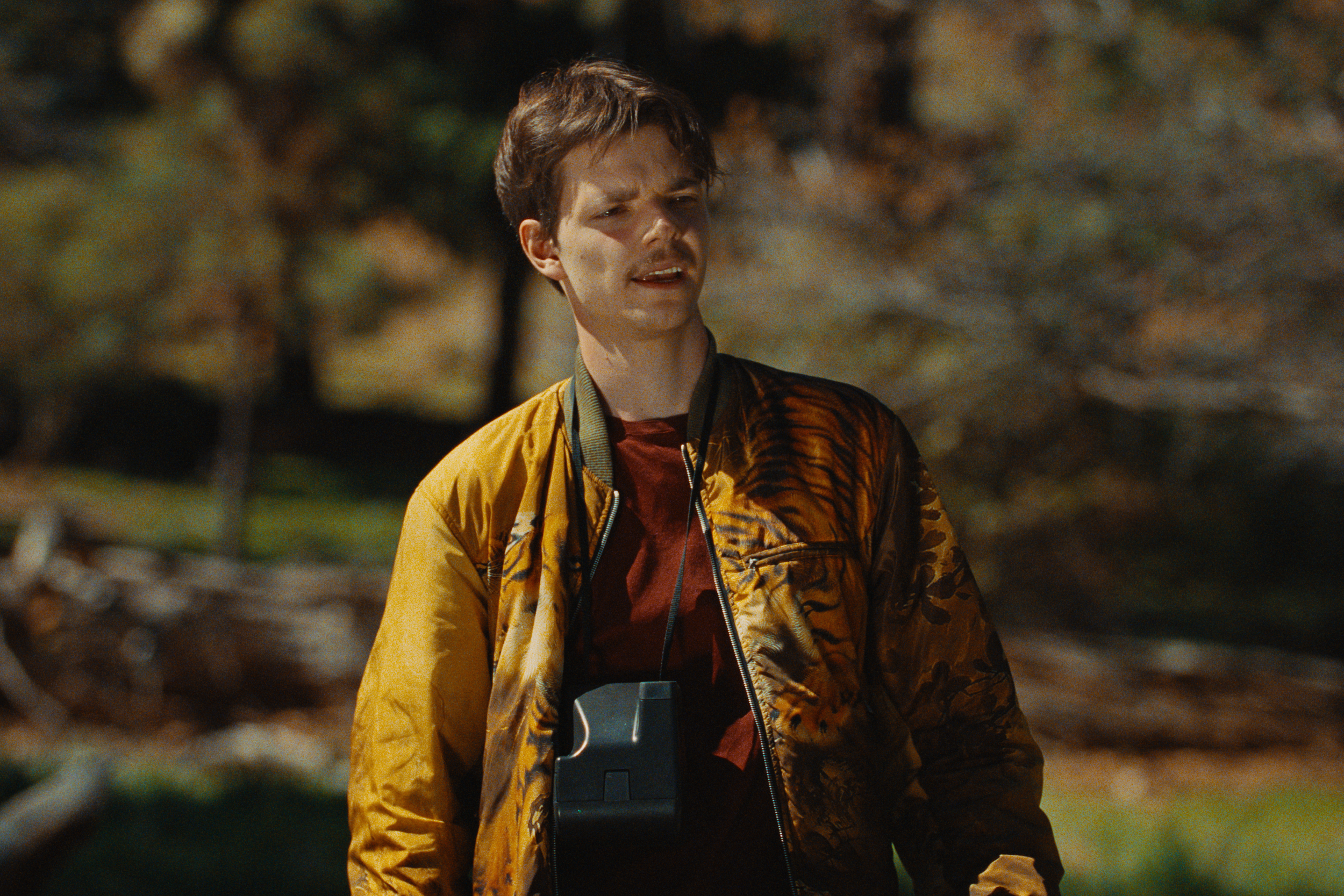 A young man outdoors in a park-like setting with autumn trees in the background. He has short dark hair, a light mustache, and is wearing a tiger print jacket over a red shirt. He has a camera around his neck and looks to the side.
