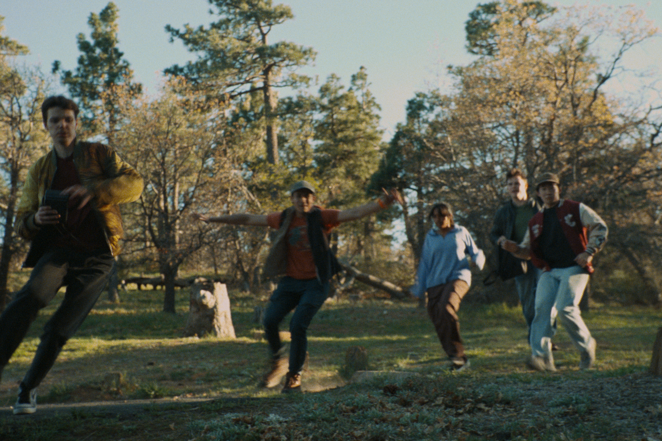 A group of five young people walking and enjoying themselves in a park with tall trees in the background during daytime.