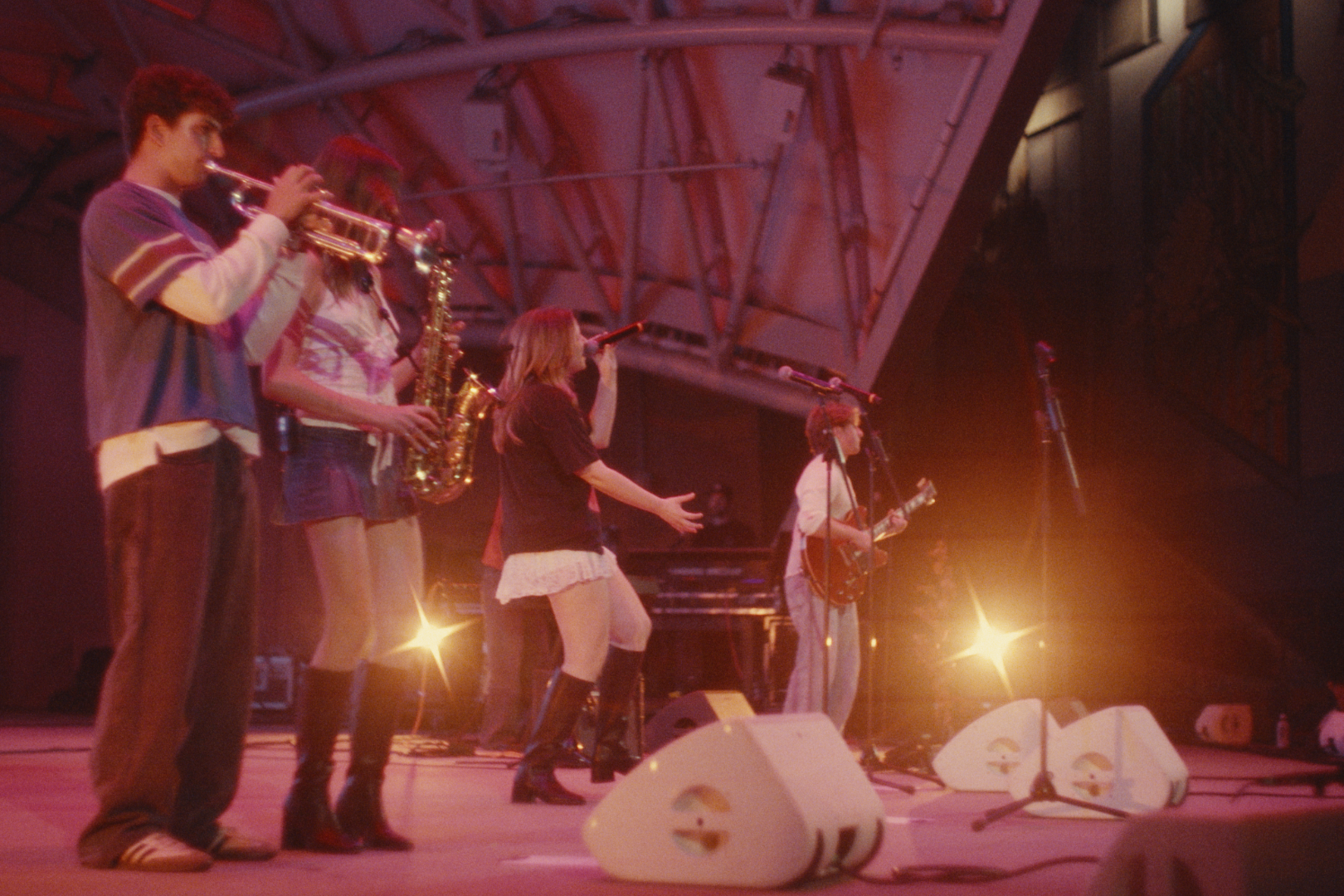 A group of five young people performing on stage, with some playing musical instruments and others singing, under colorful stage lighting.