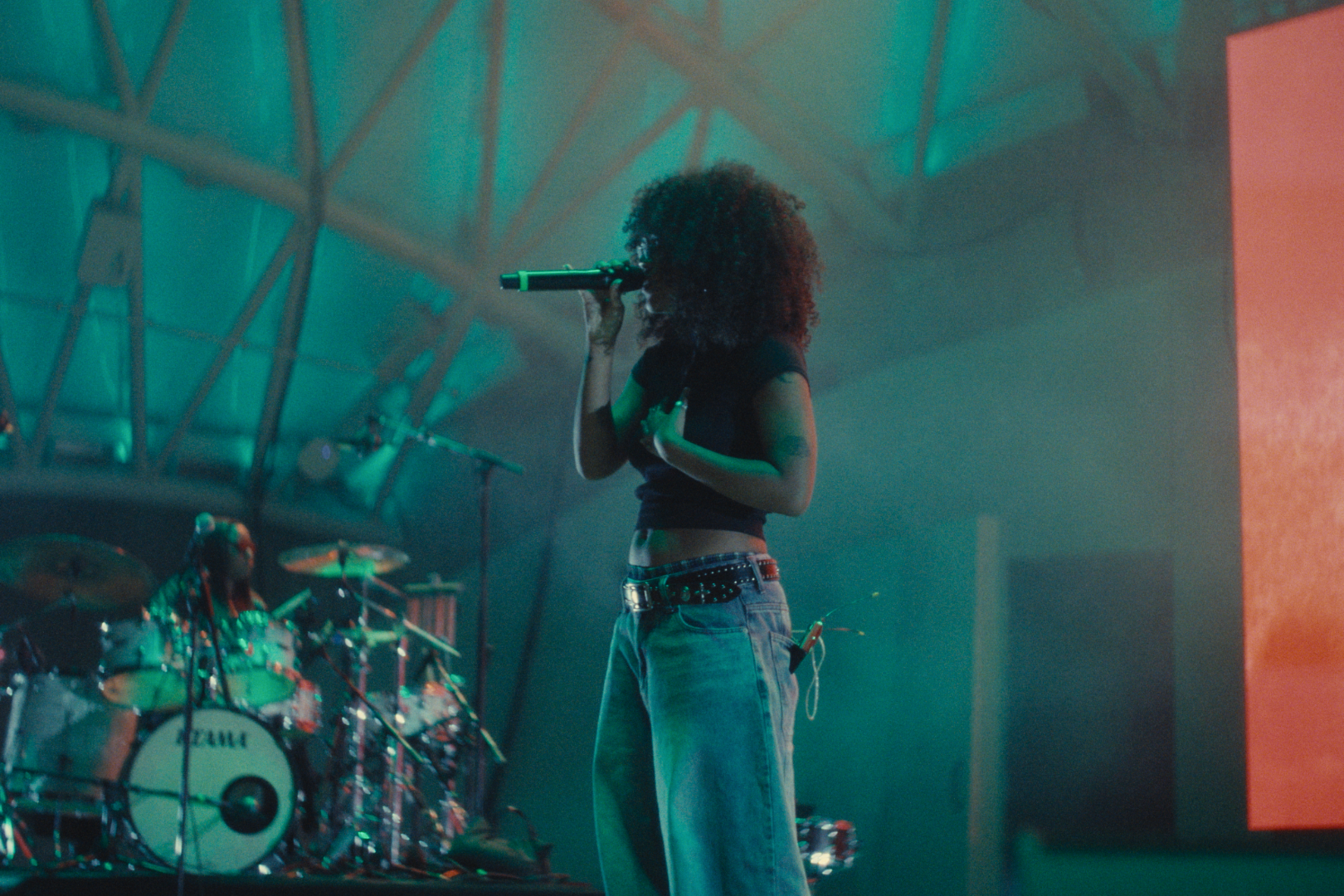 A woman with curly hair singing into a microphone on stage, accompanied by a drummer in the background. The stage is lit with green and blue lights.