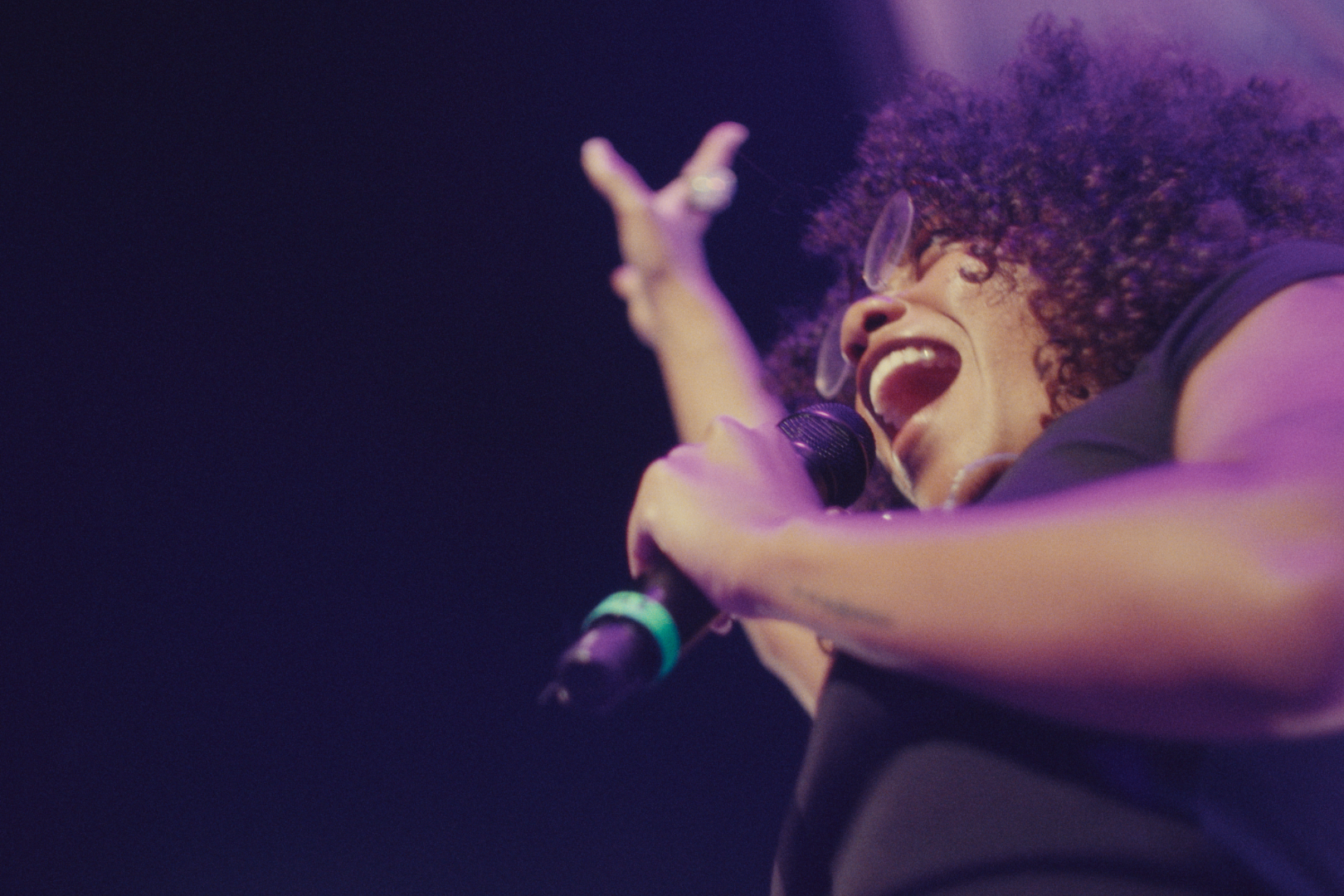 Woman with curly hair and glasses singing passionately into a microphone on stage with purple lighting.