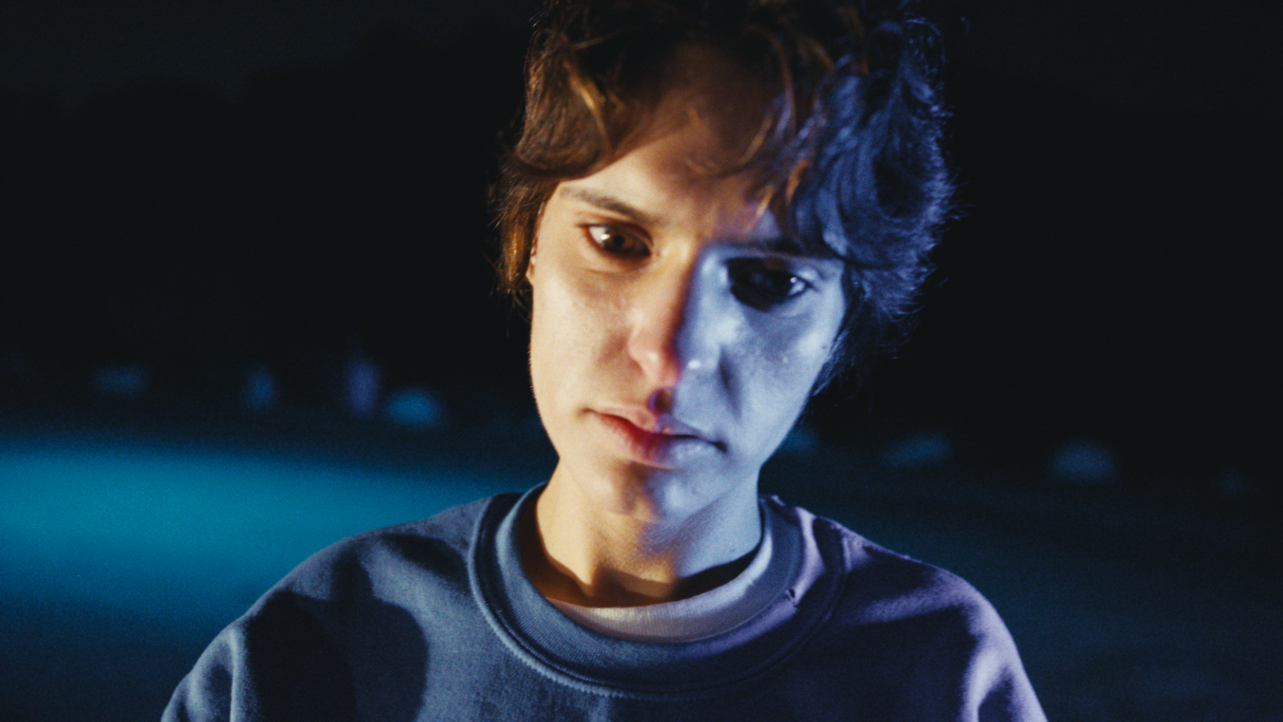 A teenage boy with brown hair and brown eyes looking downward with a neutral expression, wearing a dark sweatshirt, set against a dark background with blue lighting.
