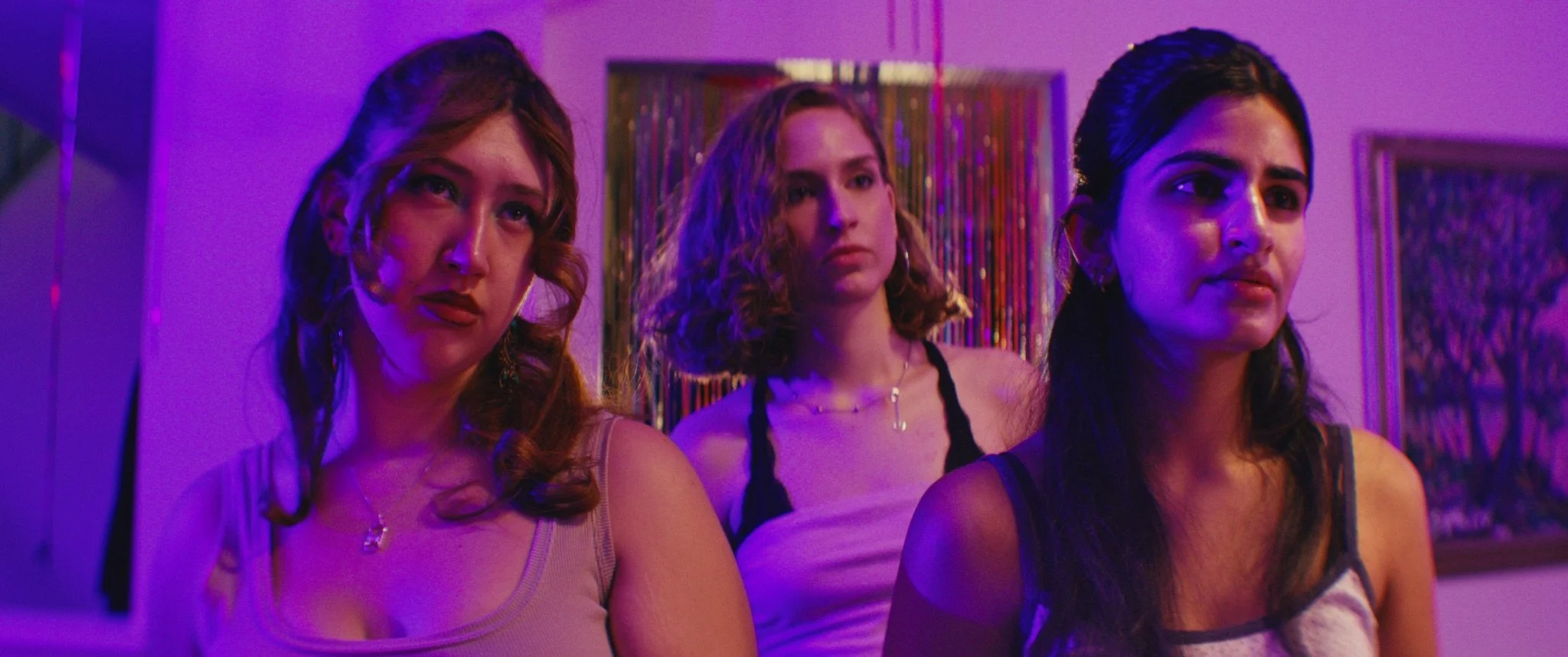 Three women standing in a room with colorful, artistic wall decorations, illuminated by blue and purple lighting.