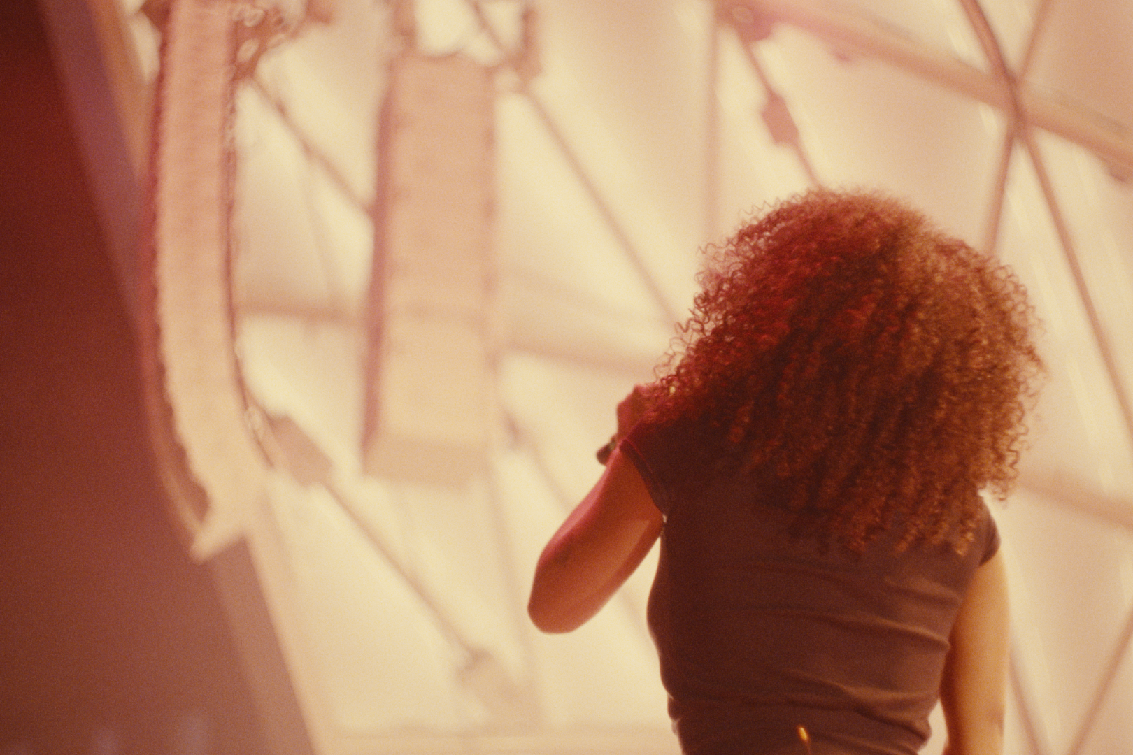A woman with curly hair standing against a large architectural structure with geometric lines and a translucent canopy.